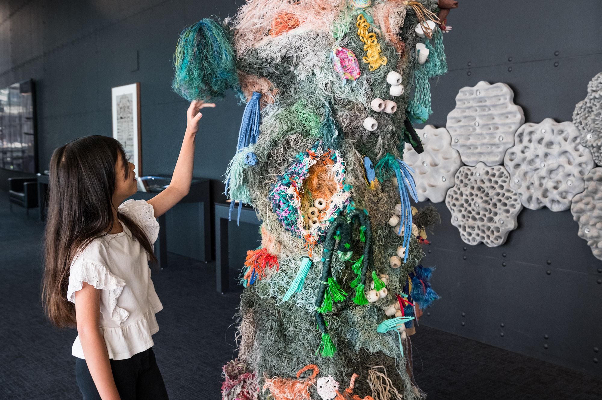 Photo of a girl with long brown hair and a white top touching a tall sculpture made from colourful recycled material to look like a coral reef.
