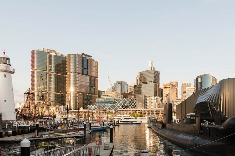 Photo of sydney skyline with a lighthouse and tall ship on the left, and a submarine on the right.