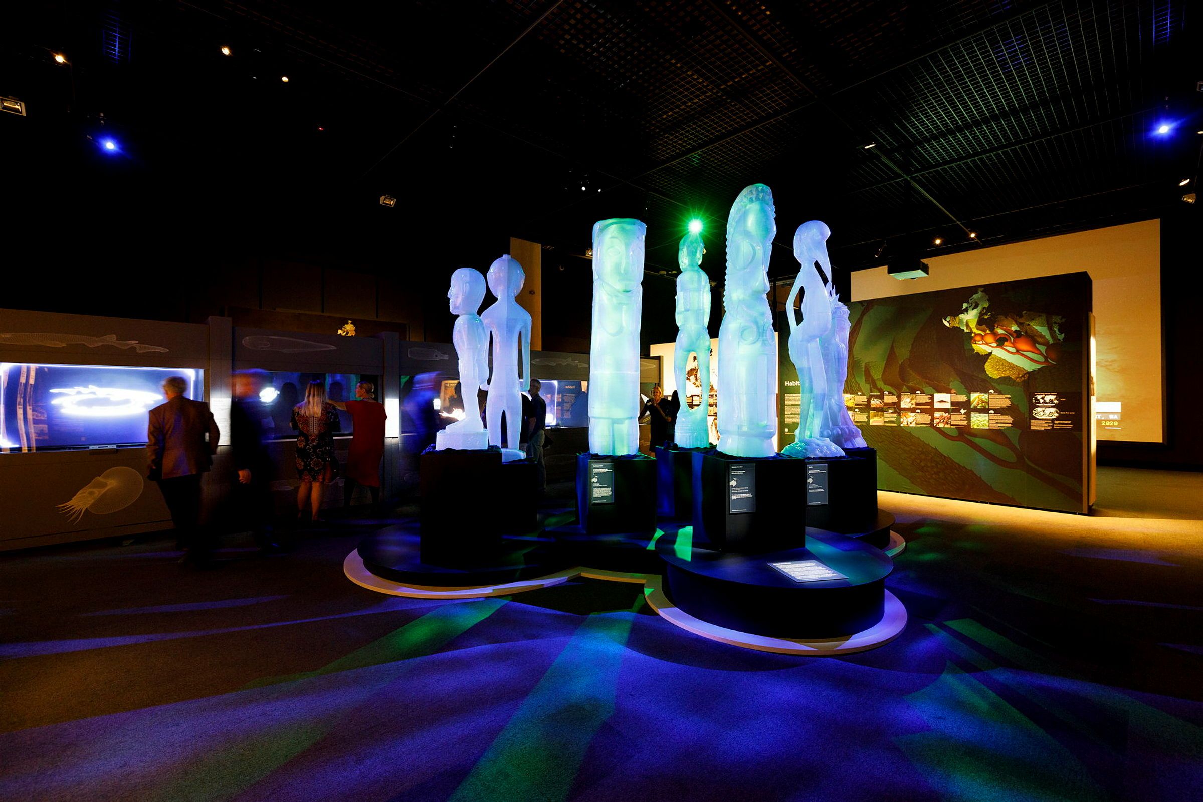 A photo of a dark museum exhibition with tall, white sculptures at centre