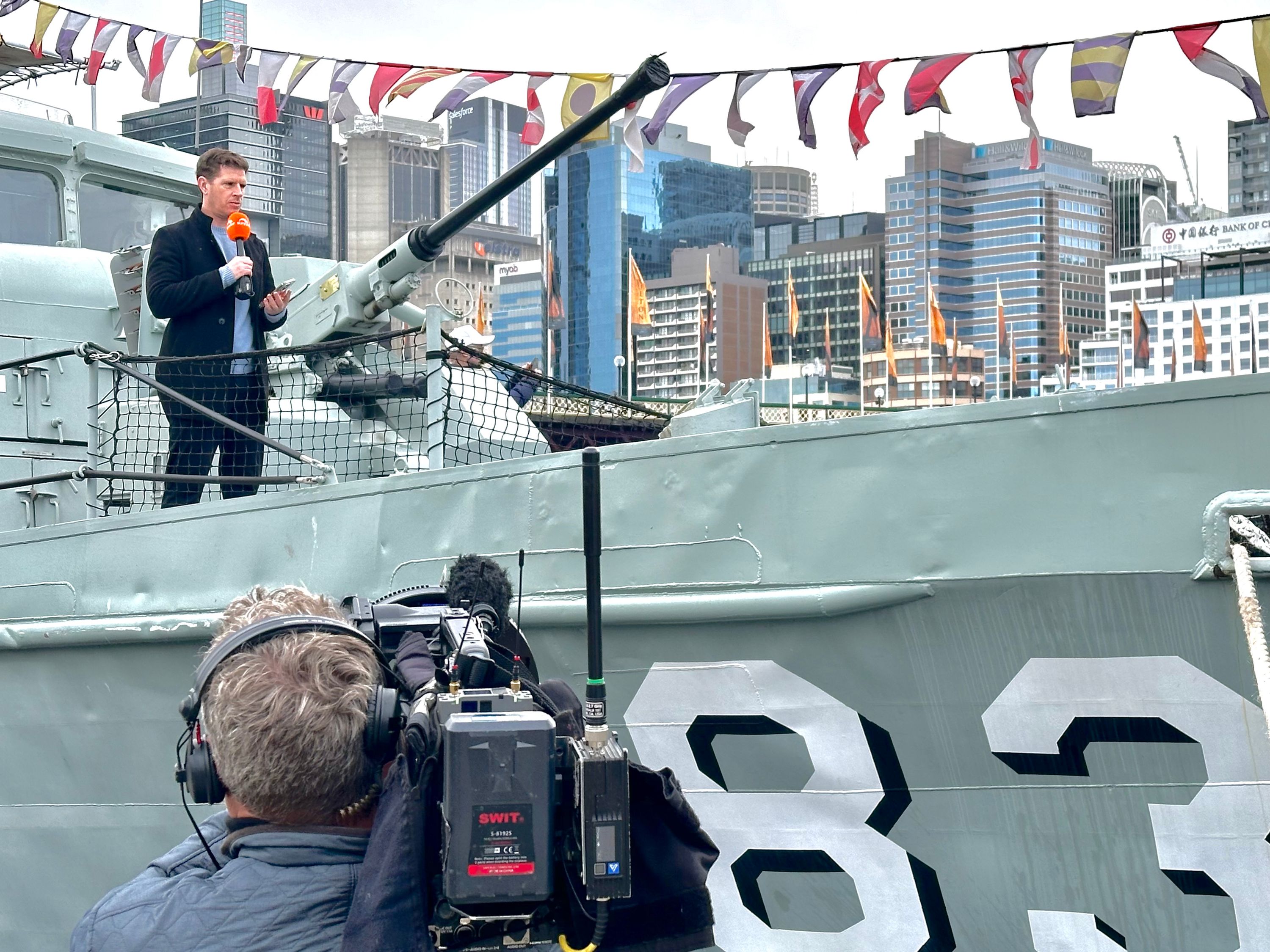 Photo showing behind the scenes off a TV shoot on a navy vessel, with a camera man in the foreground, and a male presenter standing on the boat behind.