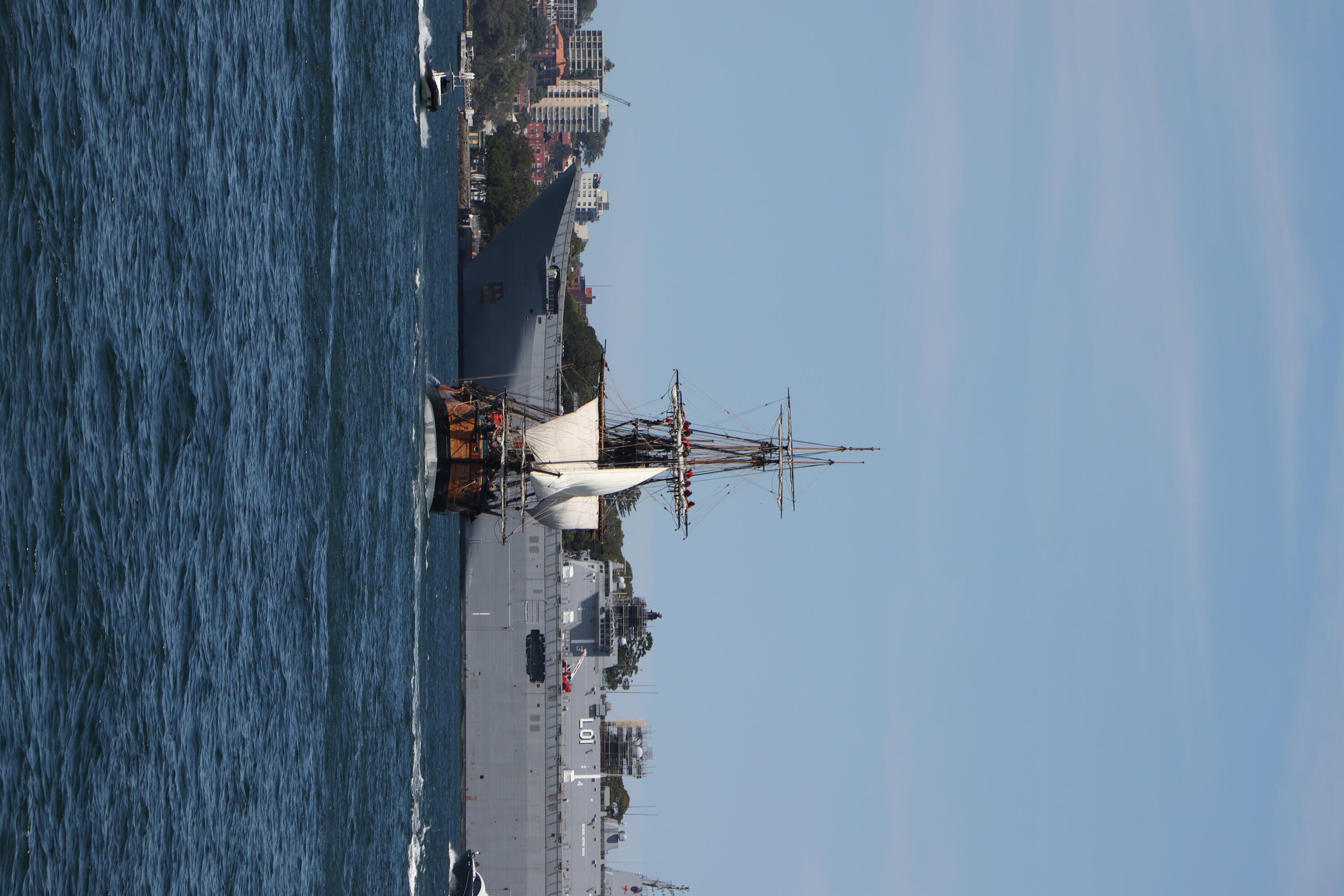 Photo showing a tall ship, Endeavour, with 2 sails up, sailing in Sydney Harbour, with part of a large Navy ship behind it. 