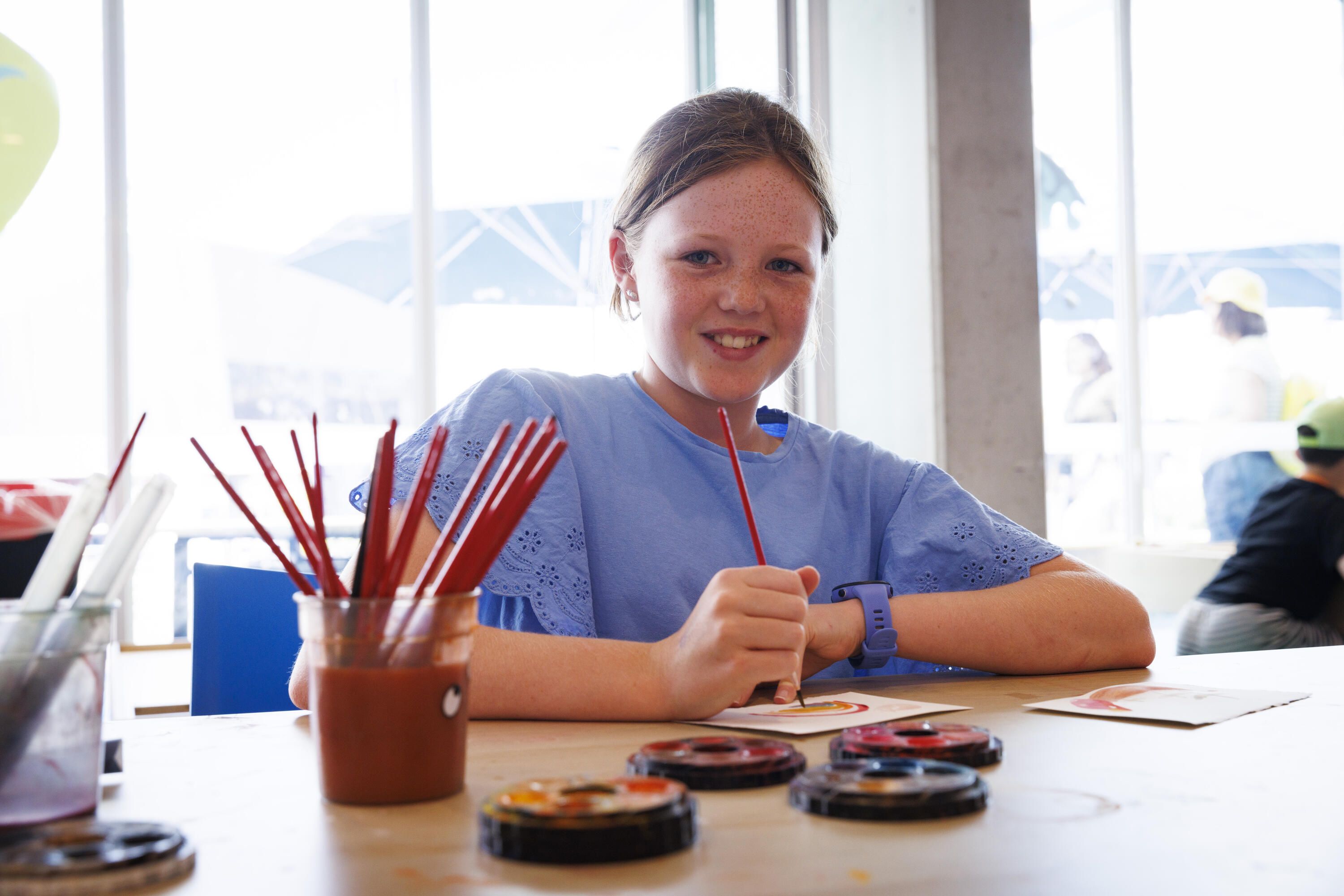 Photo of a girl wearing blue smiling at the camera while watercolour painting.