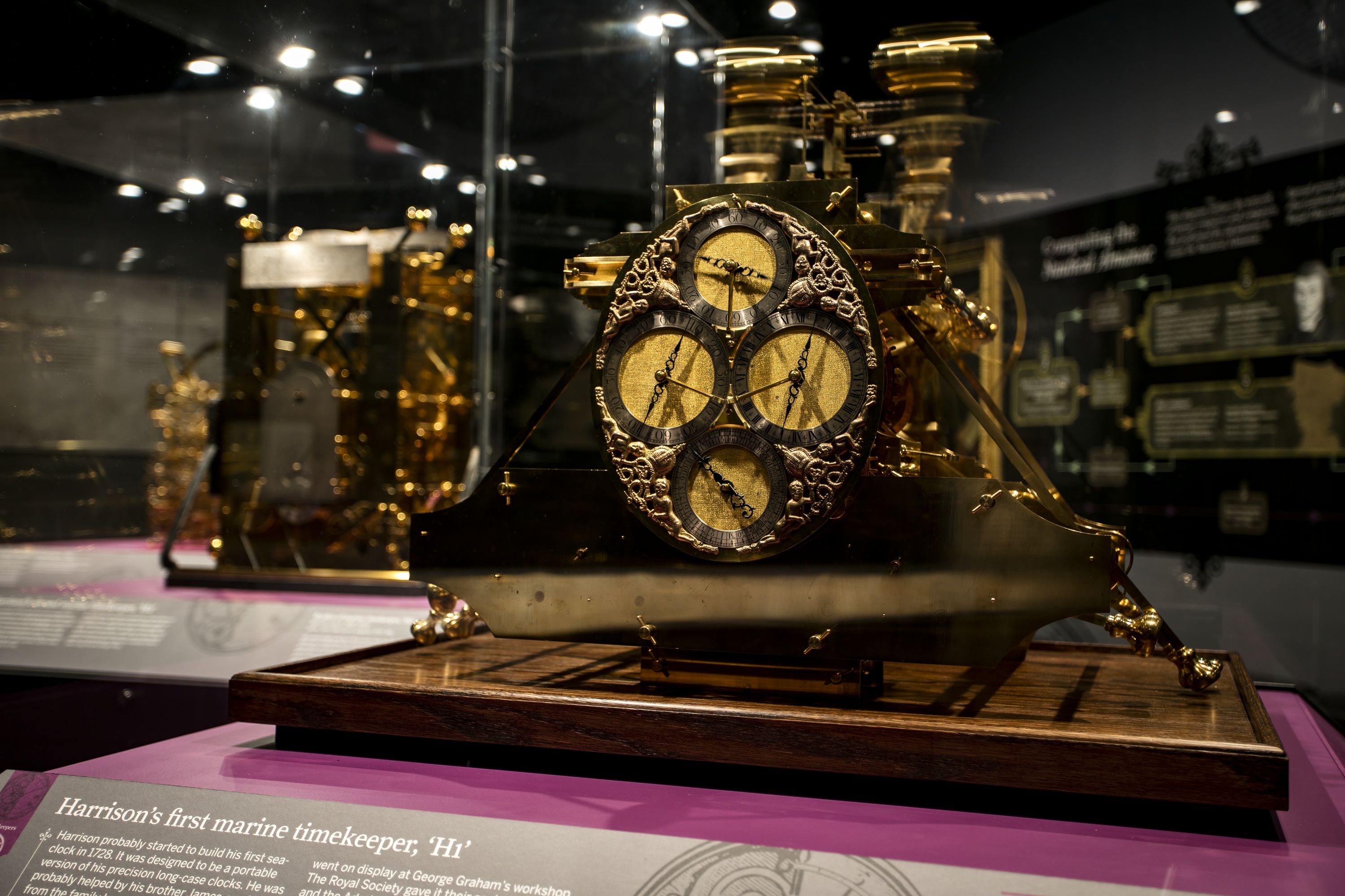 Photograph of a museum exhibition with a display case featuring an old, gold coloured clock with 4 dials. 