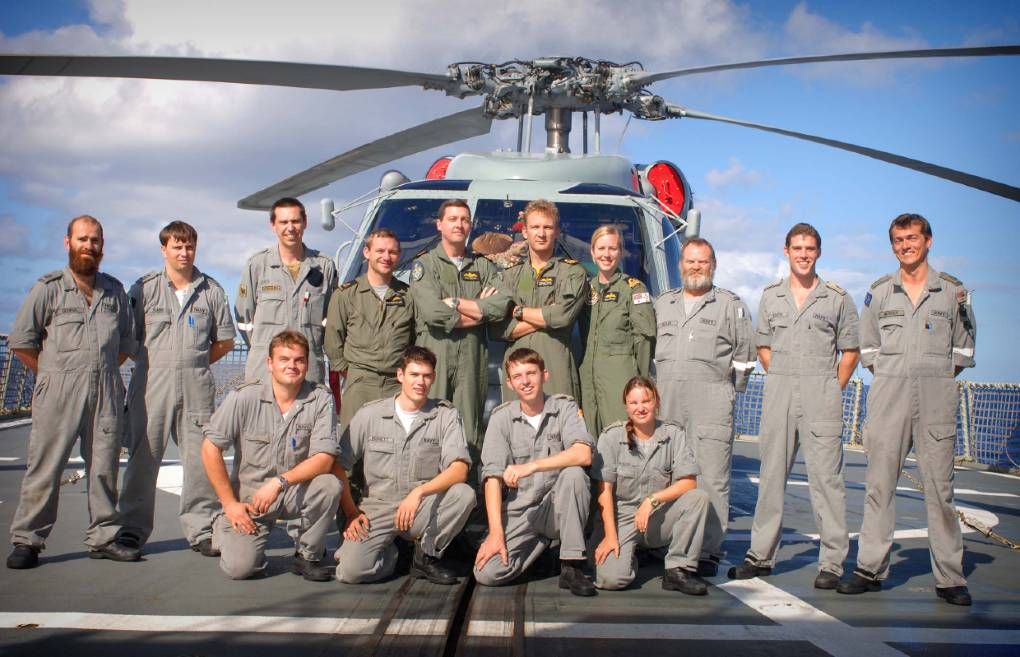 A group of military personel in uniform. The photo is taken outside standingg or kneeling infront of a grey navy helicopter.