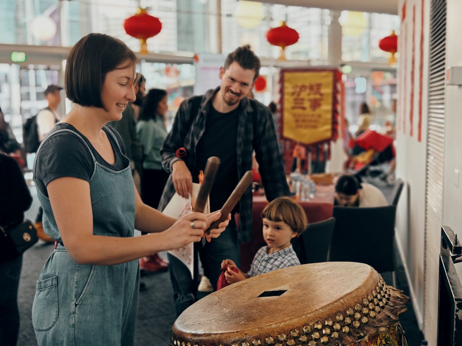 Photo of a woman with short, dark hair, wearing blue overalls and a black t-shirt, standing next to a large drum and holding 2 drumsticks. A man and a child are watching from behind the drum.