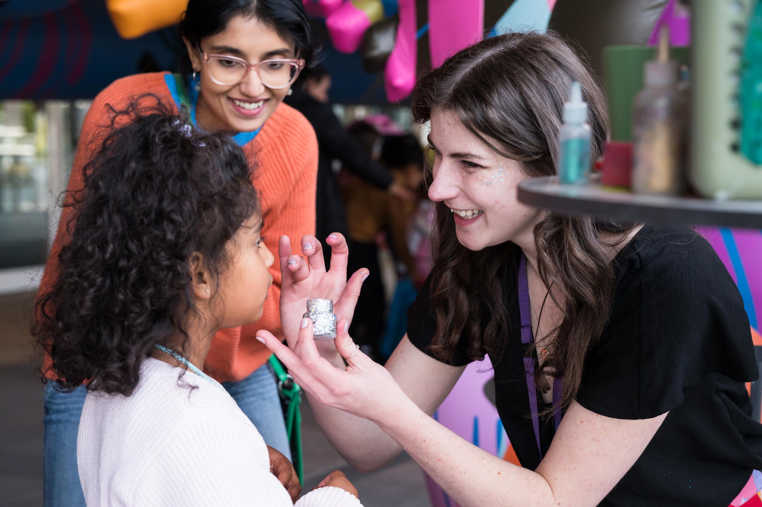 Photo of a girl getting her face painted by a young woman while her mother looks on smiling in the background.
