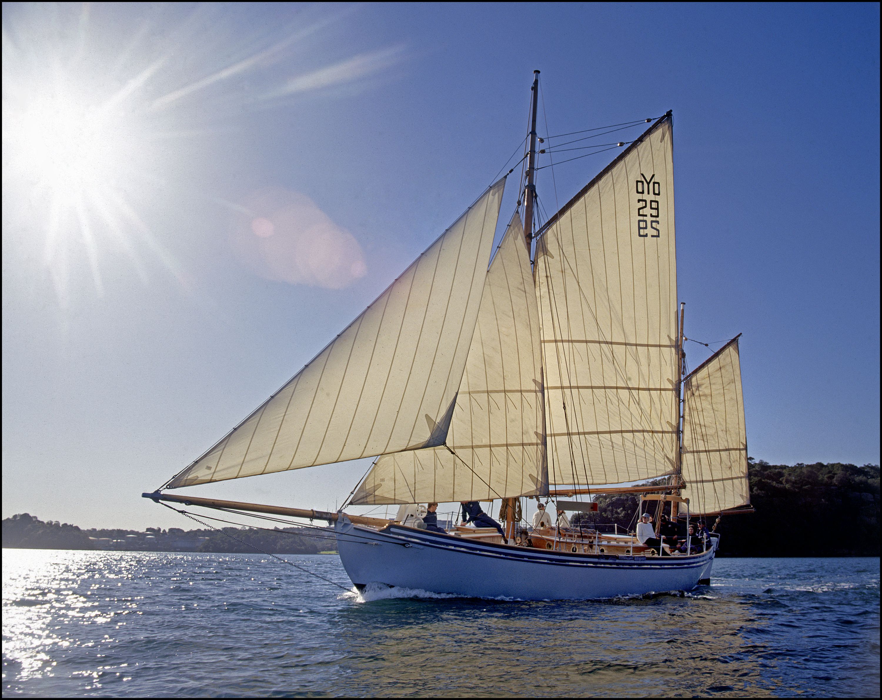 Photo of a small white boat with cream coloured sails, with lens flair from the sun behind it in the clear blue sky. 