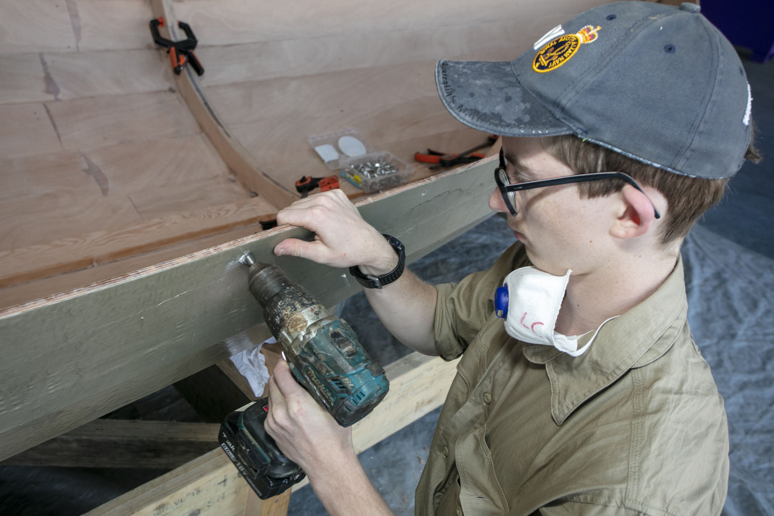 A teen boy wearing a hat and khaki shirt is using a drill to build a wooden boat. 