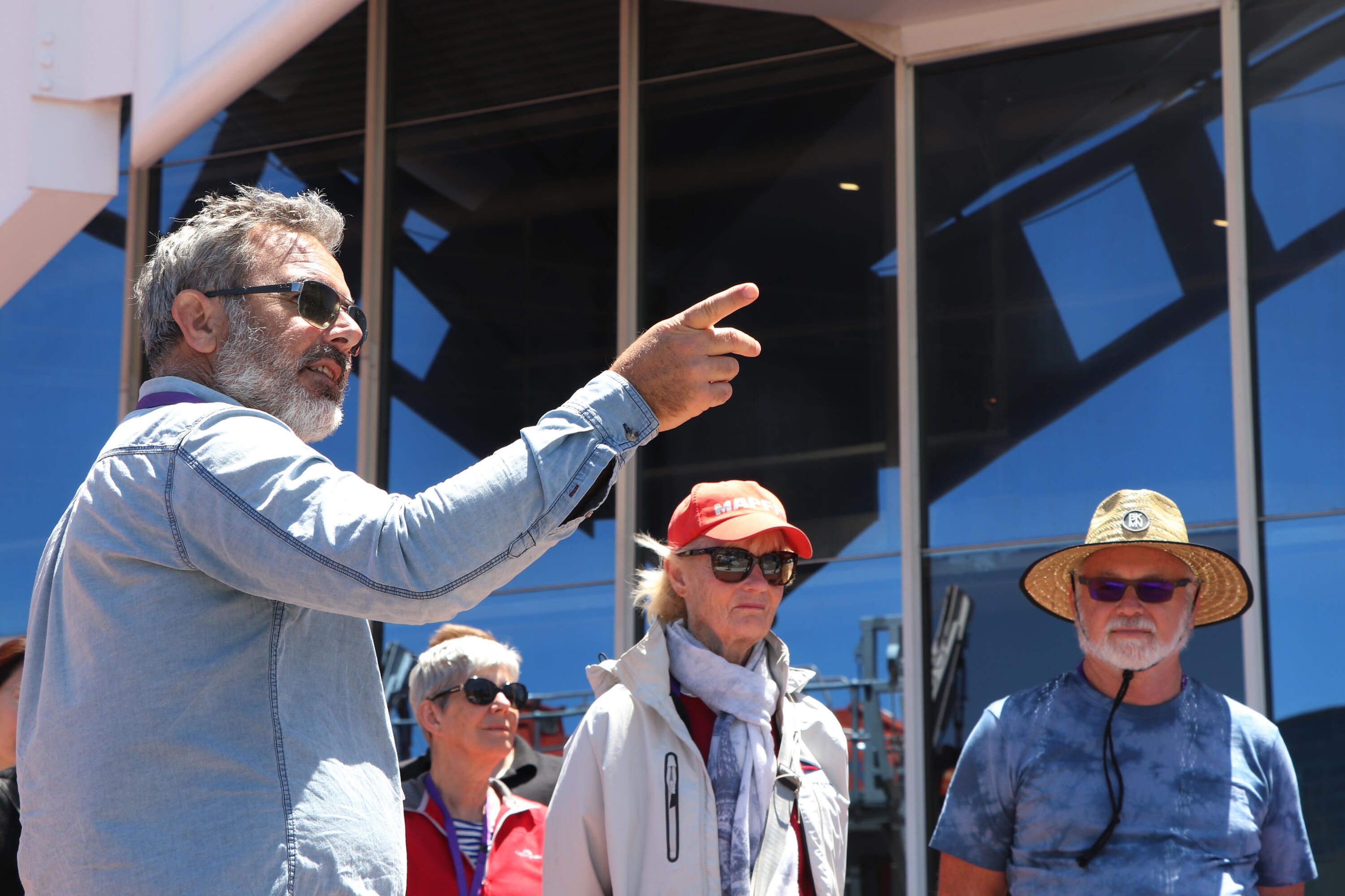 A man with grey hair and his back to the camera is pointing to the right. There are other people in the background wearing hats and sunglasses.