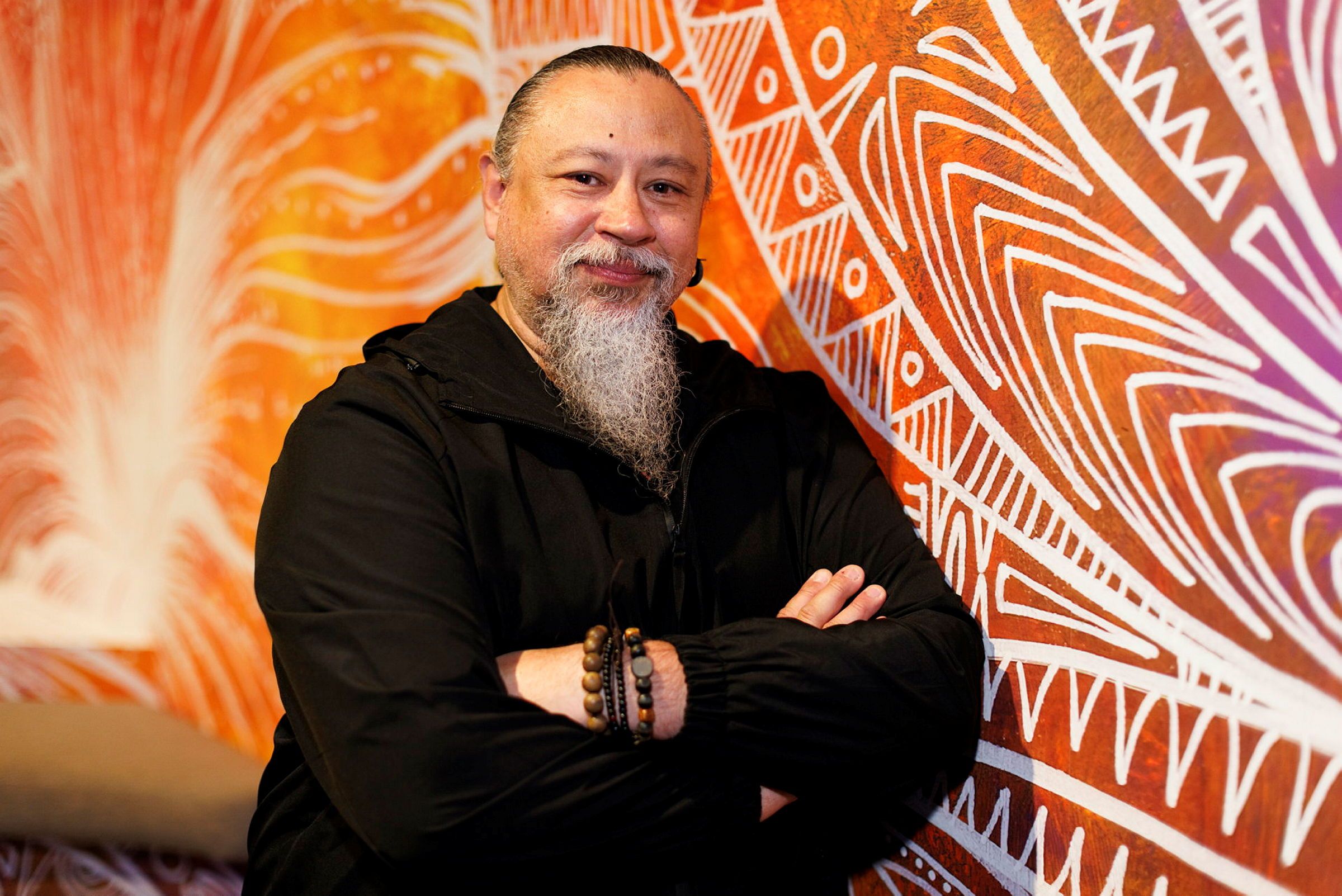 Photo of a man wearing black, with a grey beard and black hair, leaning against a wall featuring a colourful mural.