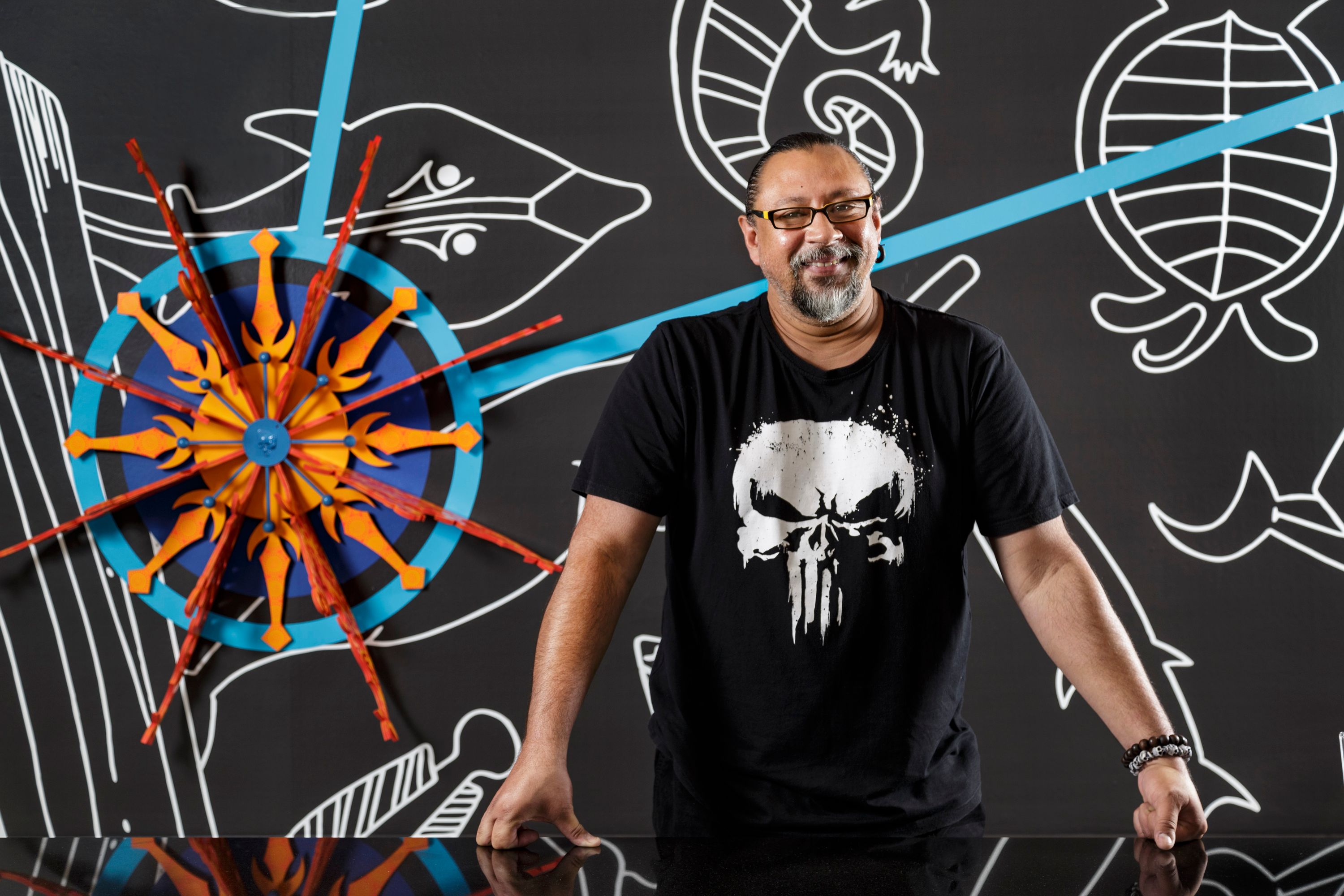 Photo of a man with dark hair pulled back, a grey beard and wearing glasses and a black t-shirt. He is standing in front of an artwork.