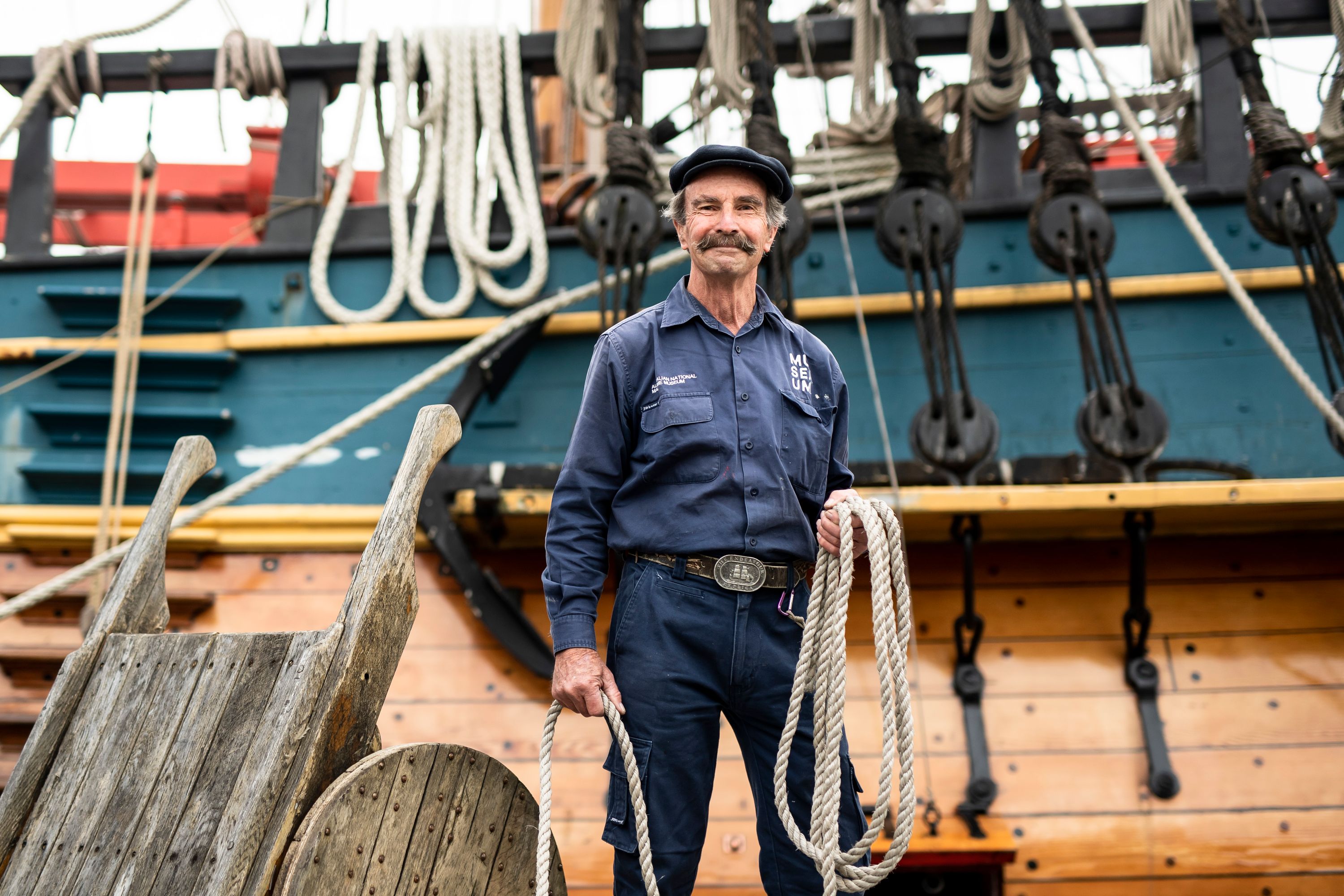 Older male Museum Volunteer standing in front of the wooden tall ship ENDEAVOUR. He holds rope coiled in his hand.