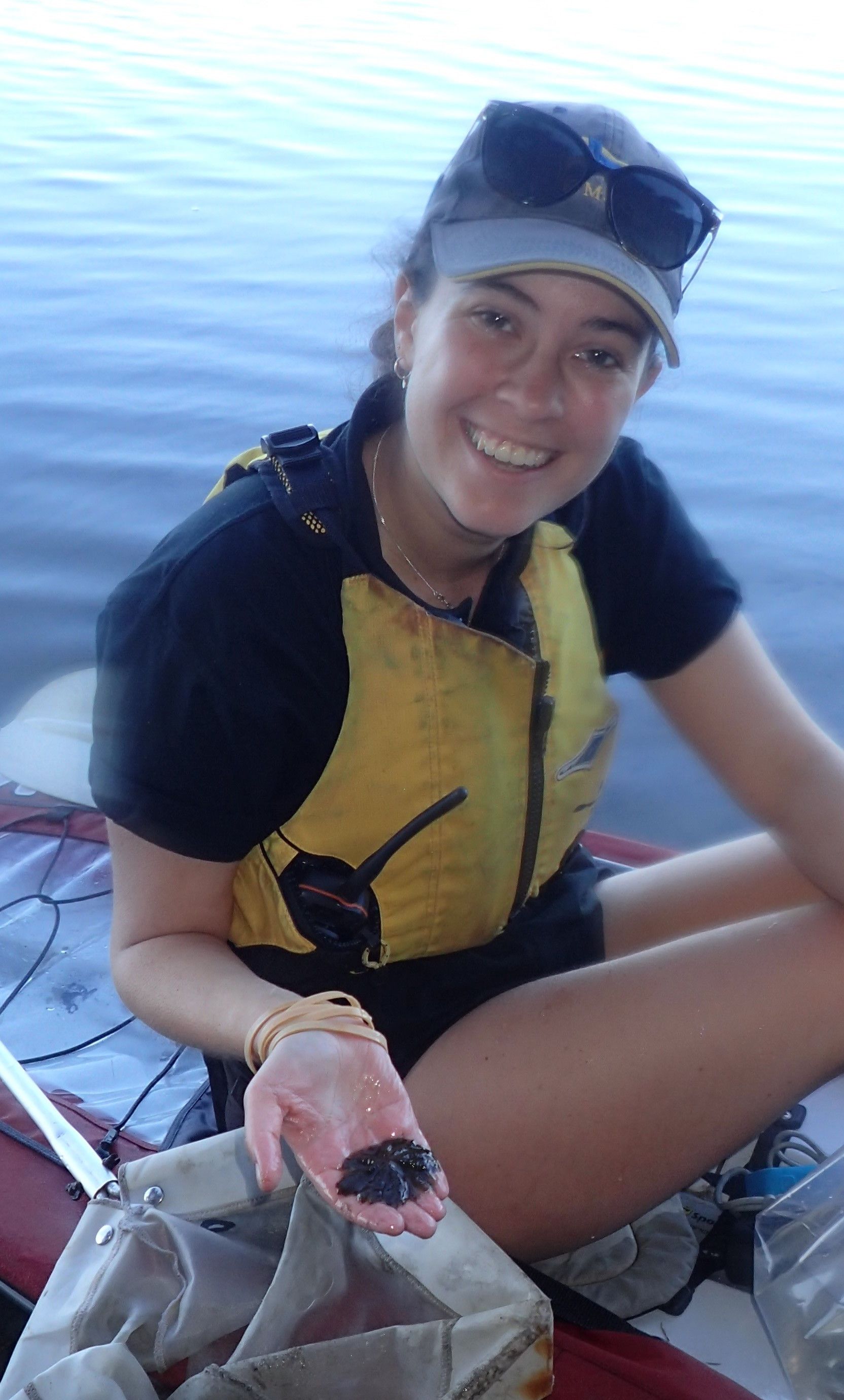 Photo of a smiling woman with a cap and sunglasses on her head, wearing a lifejacket over a black tshirt