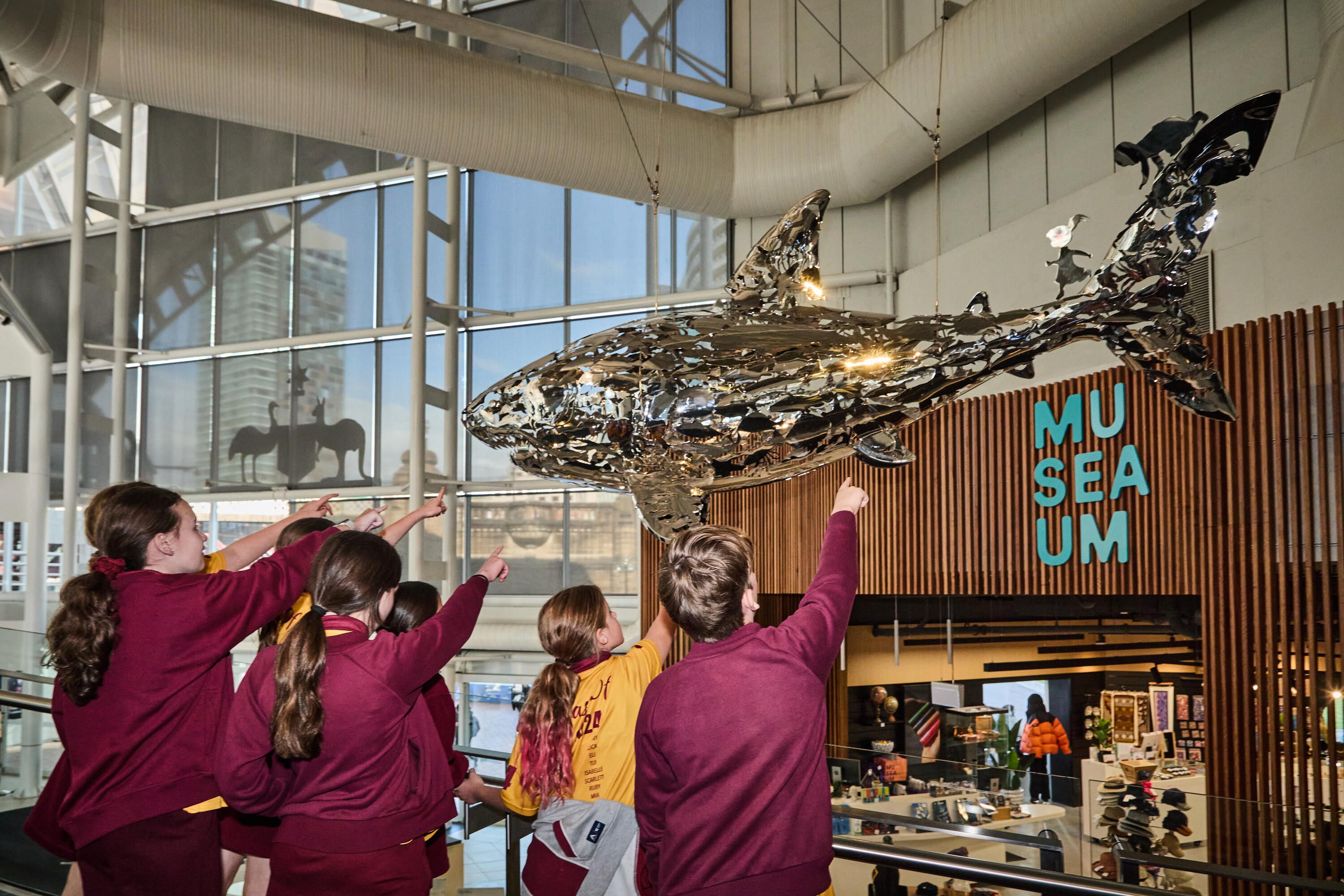 Photo taken inside the museum, with a group of students pointing up at a silver sculpture of a shark.