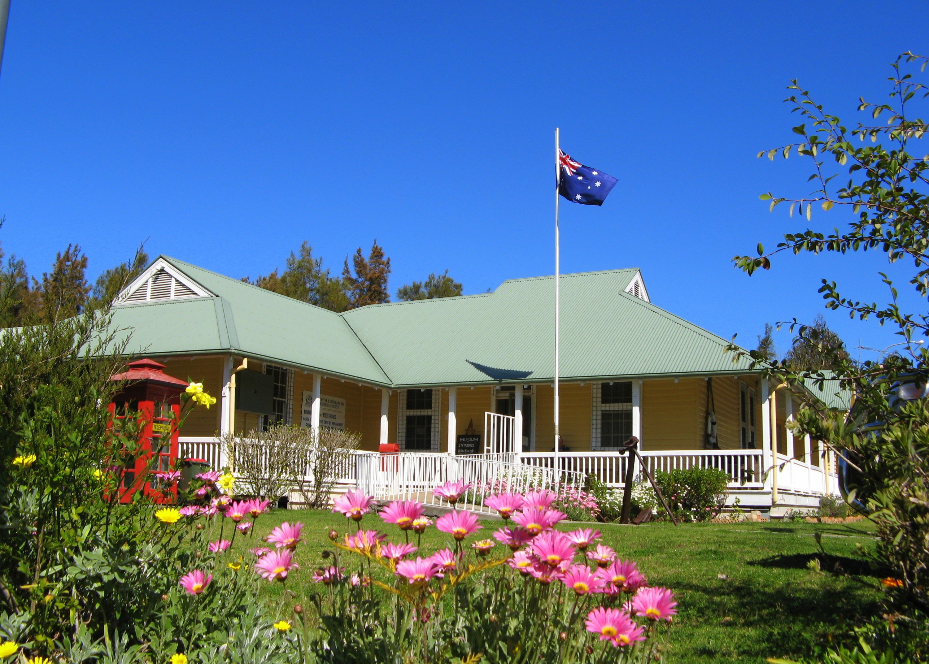 Photograph of a historic house with a green roof and veranda. There is an Australian flag on a flagpole and a garden with colourful flowers.  