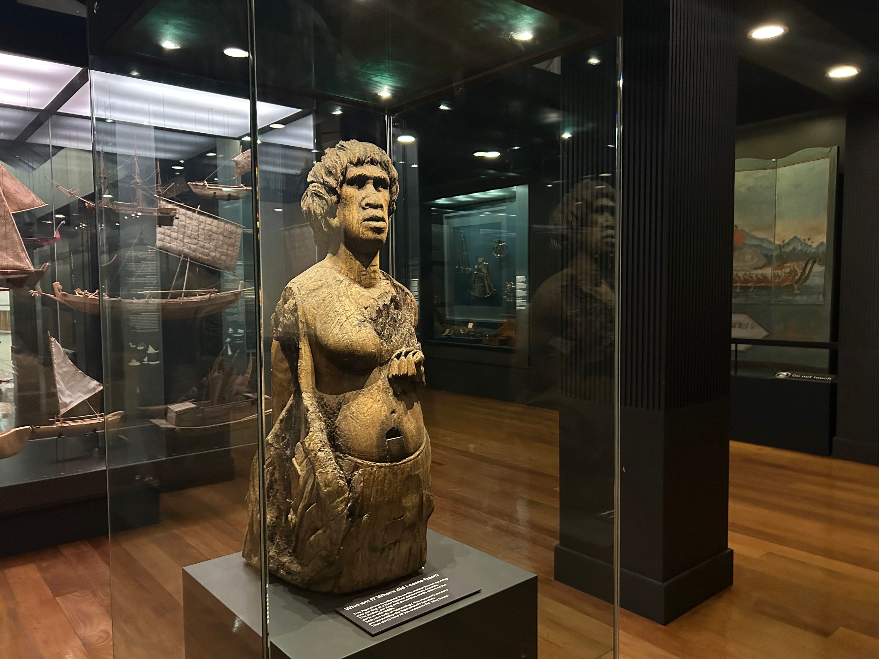 Photo of a museum gallery, with a wooden sculpture of a woman in a glass display case.