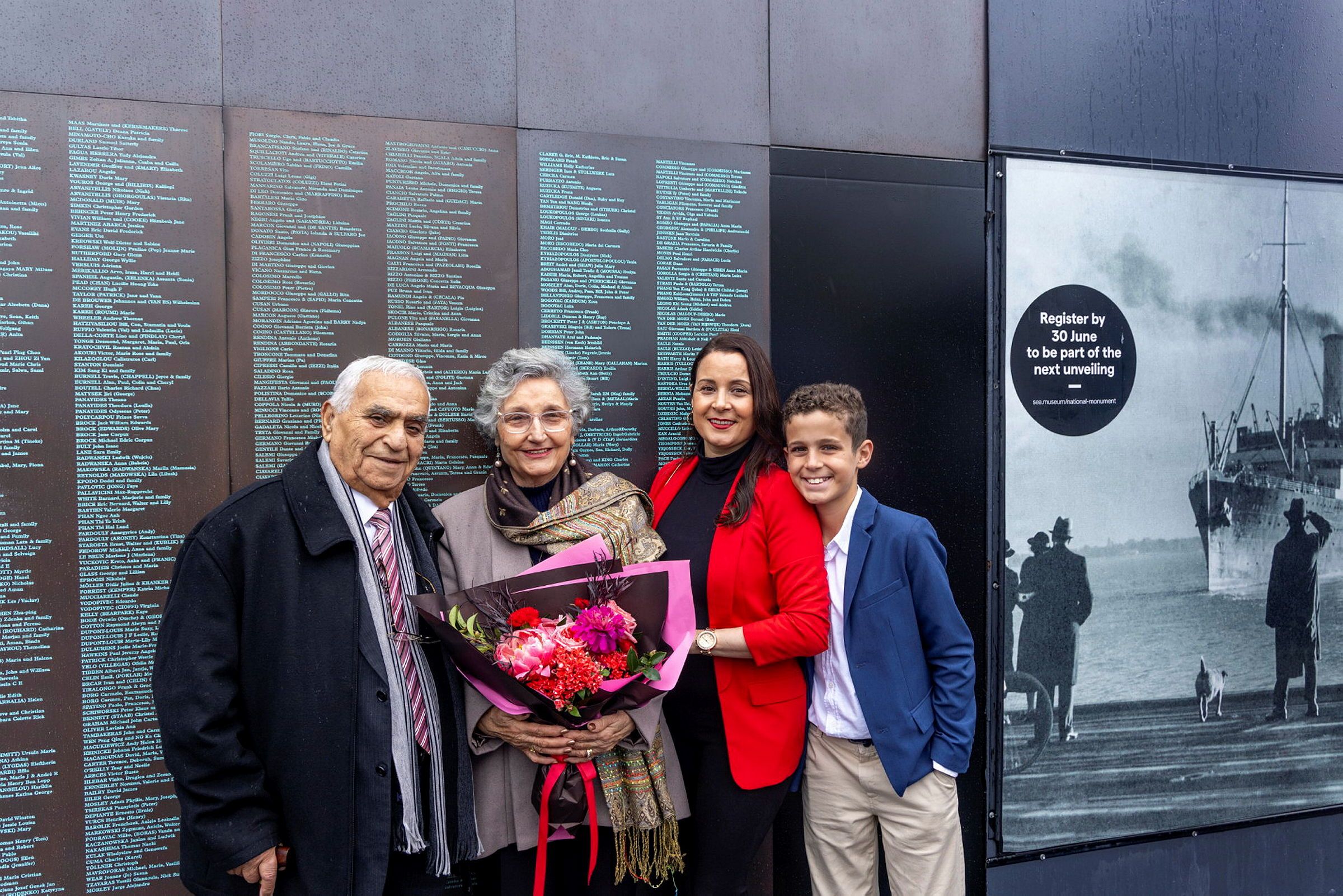 Photo showing a family; 2 grandparents, a woman and a boy standing in front of a wall with names engraved on it. 