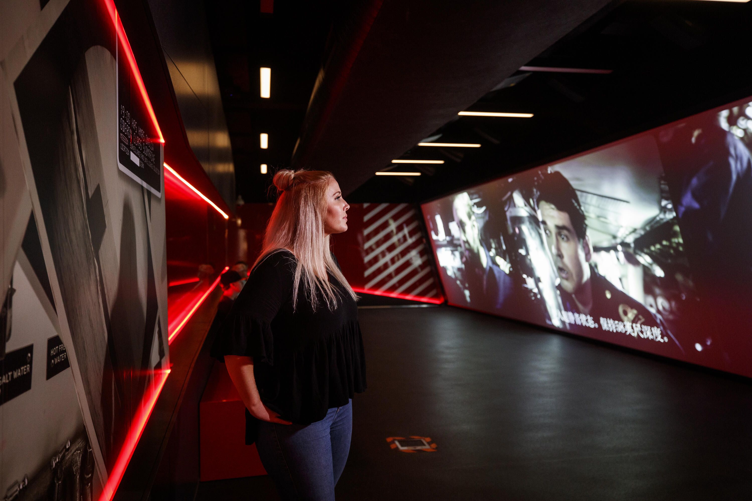 Photo of a young female adult visitor exploring a museum exhibition, watching a short film that captures the experience of the navy vessels during operations on a giant projection screen. 
