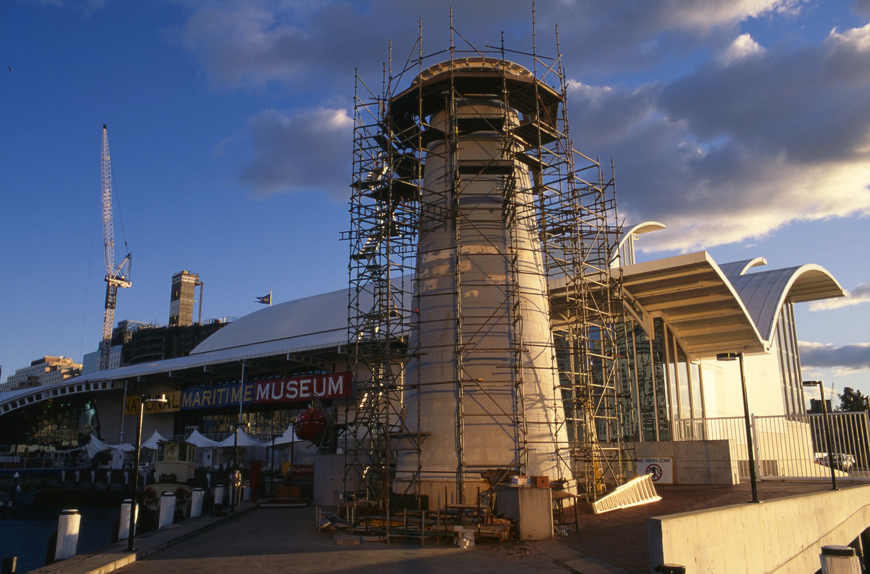 Photo of a lighthouse being constructed while surrounded by scaffolds. Photo taken at sunrise with the museum in the background and clouds overhead. 