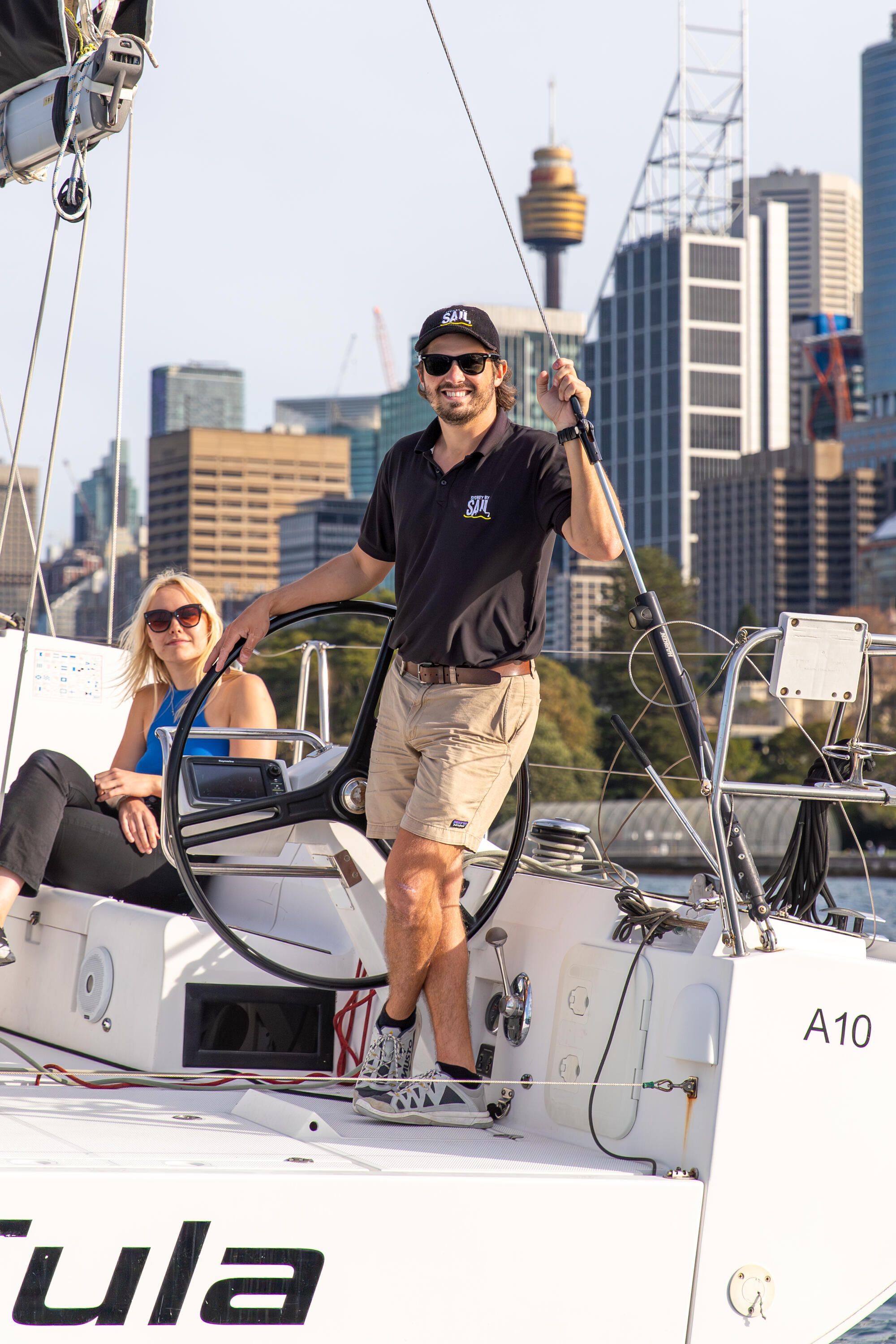A man wearing a black polo shirt, cap and sunglasses standing on a yacht. 