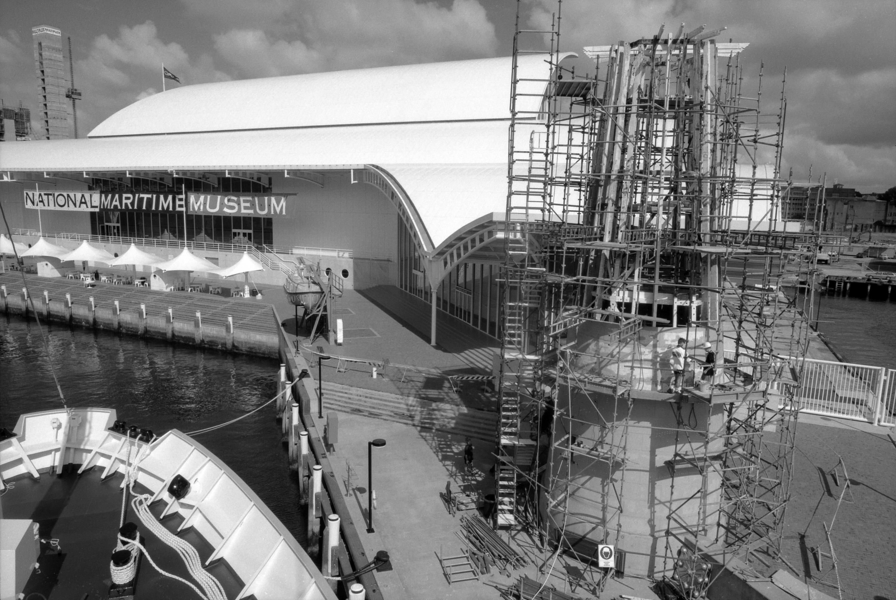 Black and White photo taken from a high elevation showing a lighthouse, covered in scaffolding, being constructed with the museum in the background. 