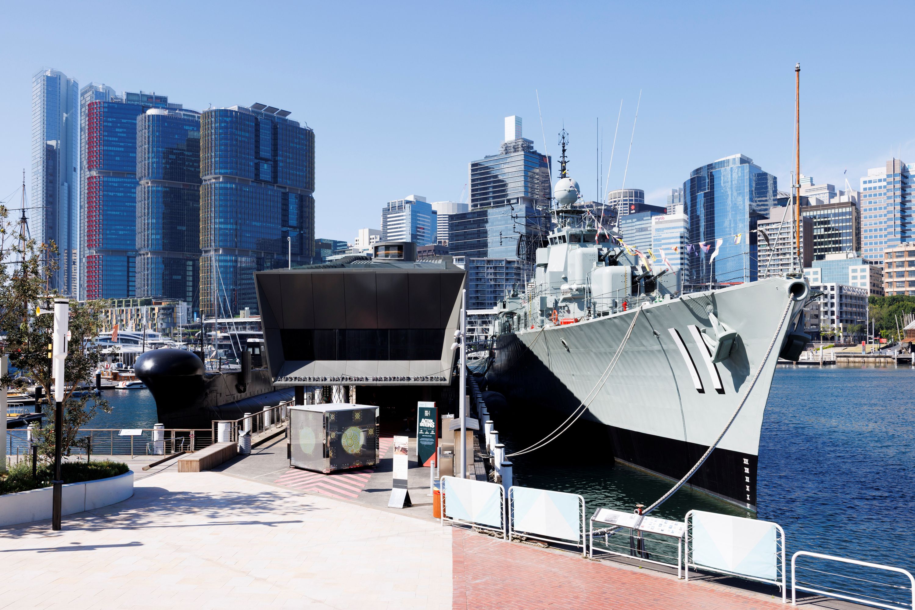 Photograph showing a building on a wharf, with a navy ship and submarine on either side.