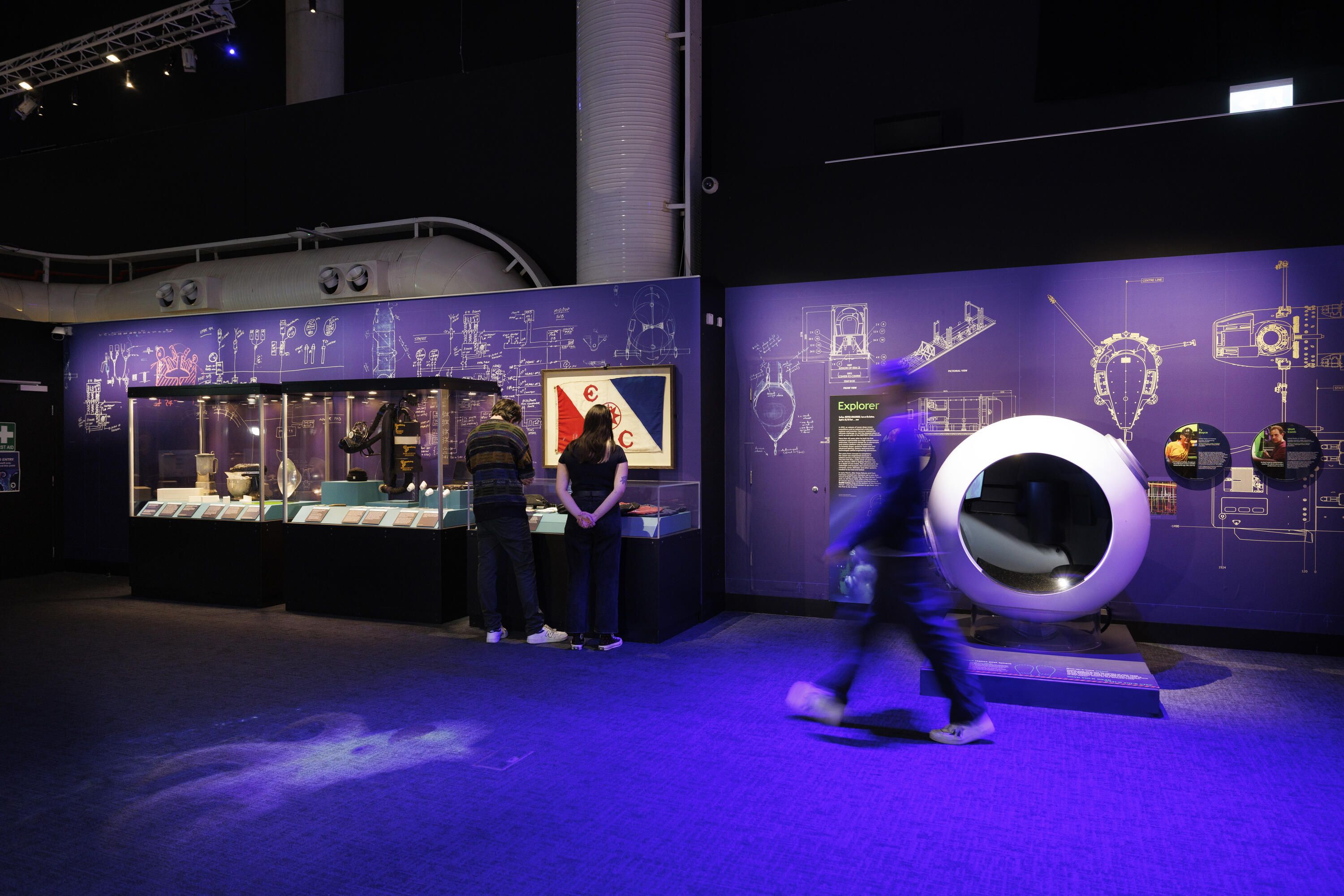 3 figures in a museum exhibition. There are display cases, blue walls with white technical drawings printed on it, and a framed flag hanging. On the right is a white spherical object.  