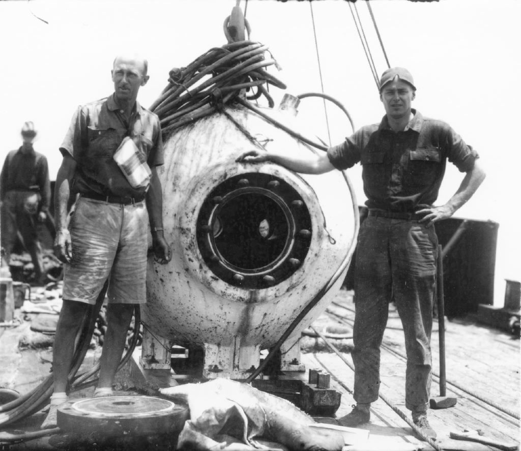Black and white photo showing 2 men next to a large sphere that comes up to their shoulder height, with a round window in the middle.