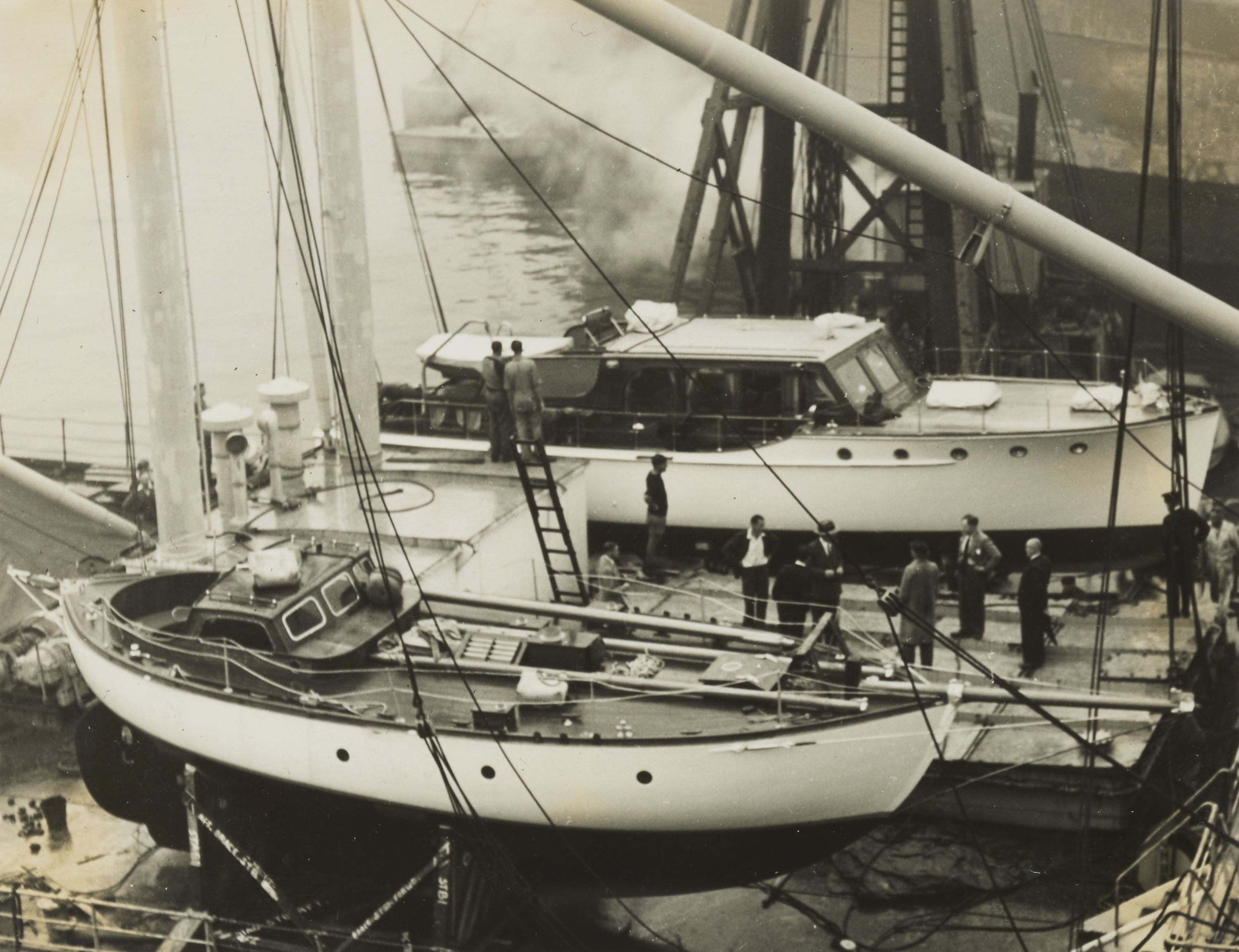 A black and white photo on yellowed paper showing a dockyard, with two boats out of the water, a sailing boat in the front and a cruiser behind it. 