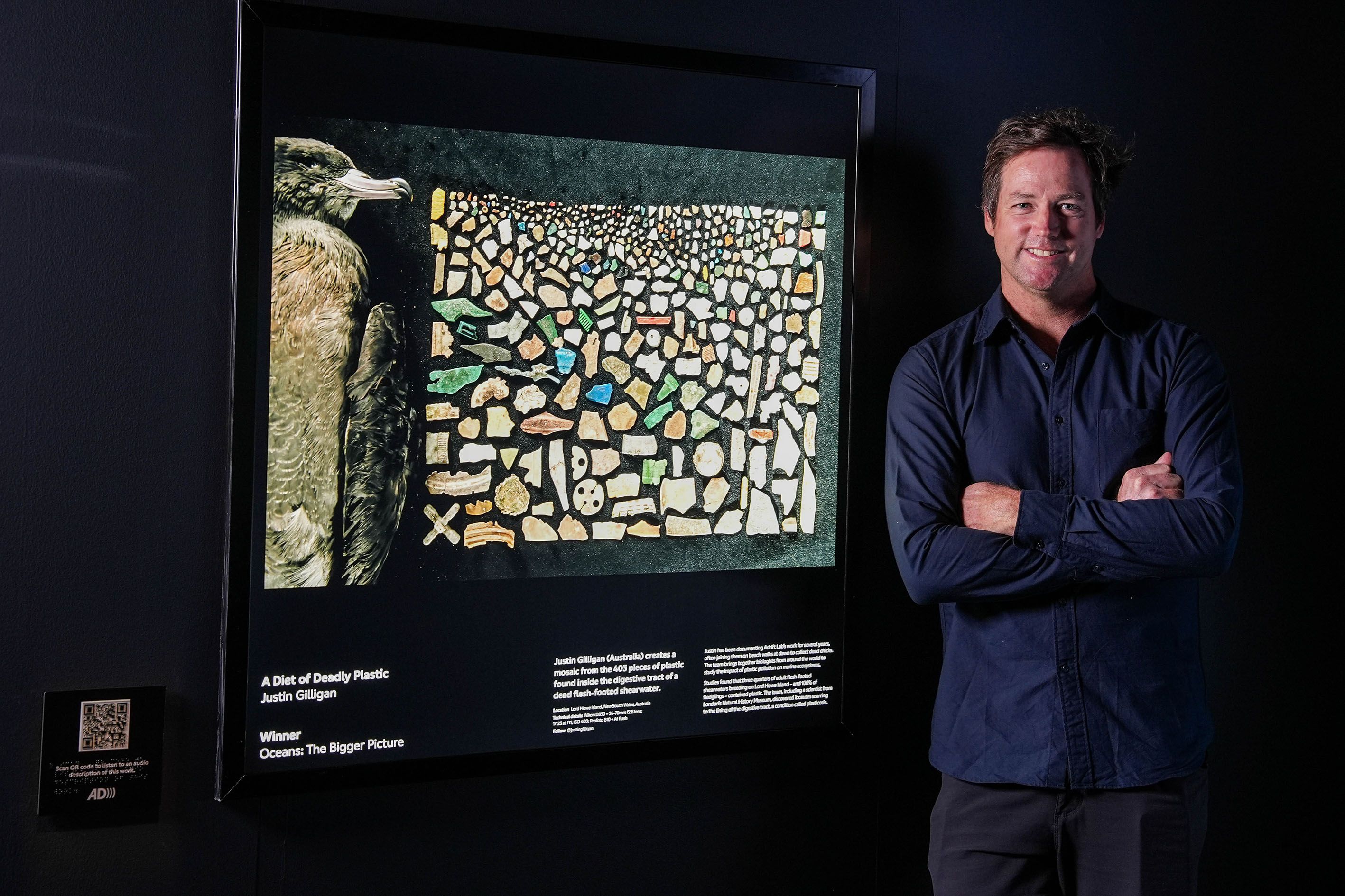 Photo taken inside a dark museum exhibition of a man beside a photograph of a dead bird and plastic.