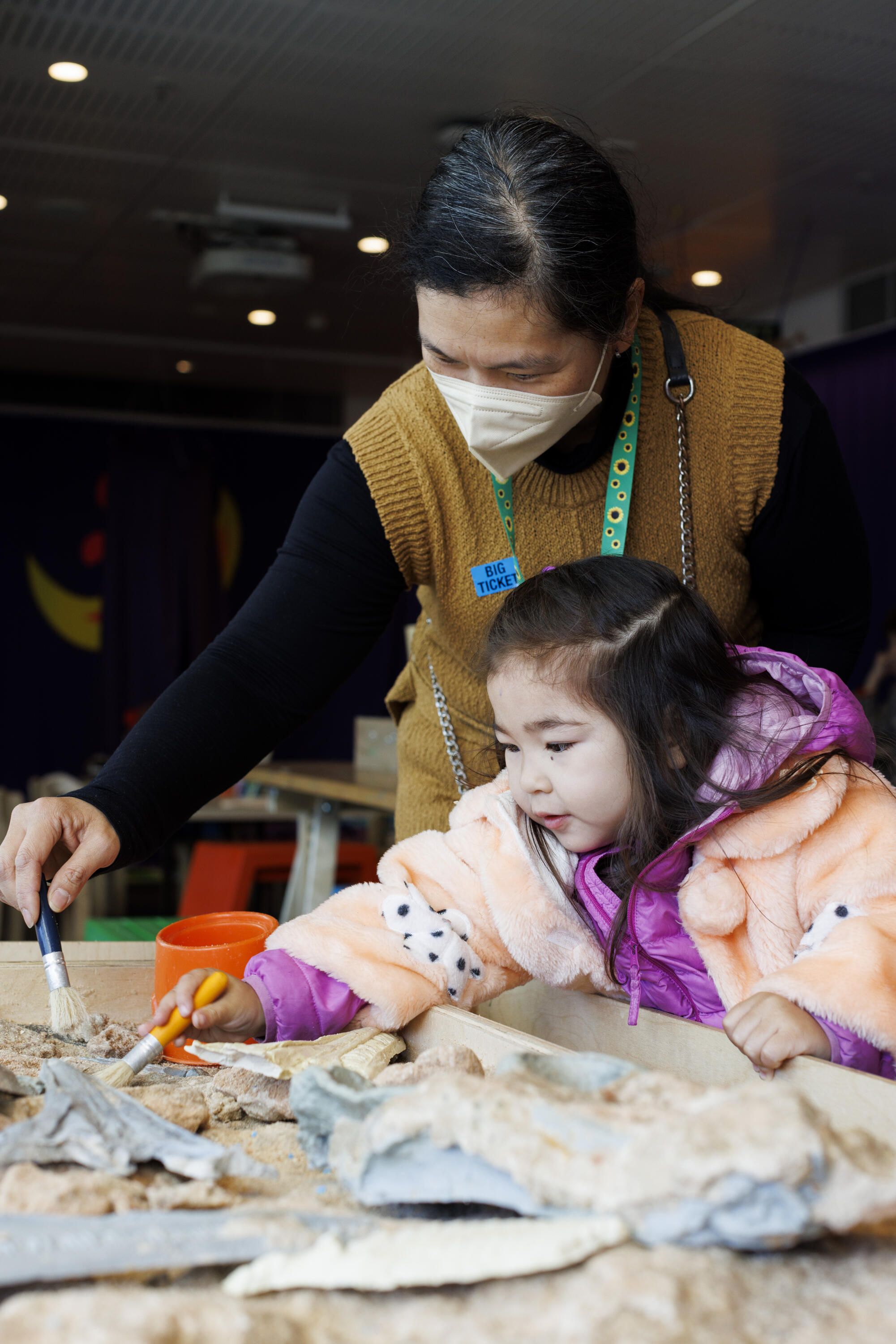 Lady with dark hair and a sunflower lanyard and holding a paintbrush, helping a young girl with an activity