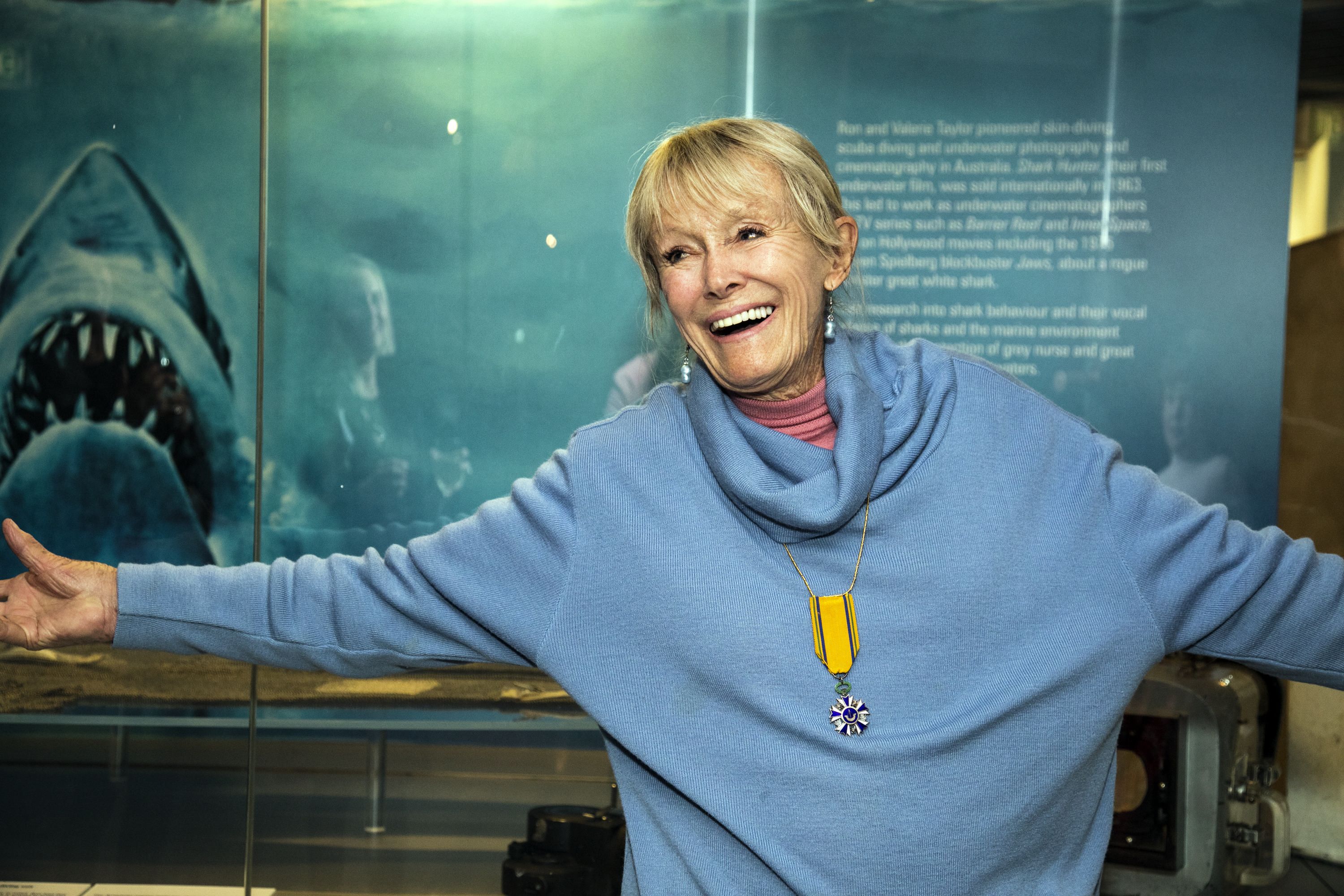Photo of an older women with blonde hair wearing a blue jumper, smiling at the camera with her arms wide.  