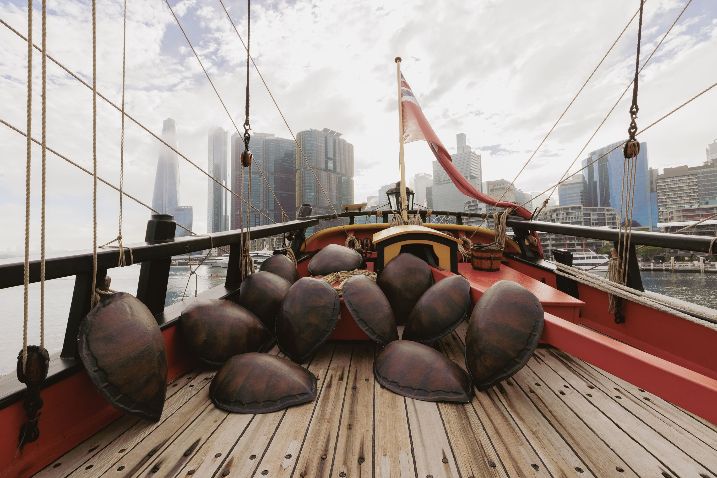 Photo showing the back of a wooden tall ship with a collection of turtle shells on the deck. There is a red flag flying on the ship, and city buildings in the background. 