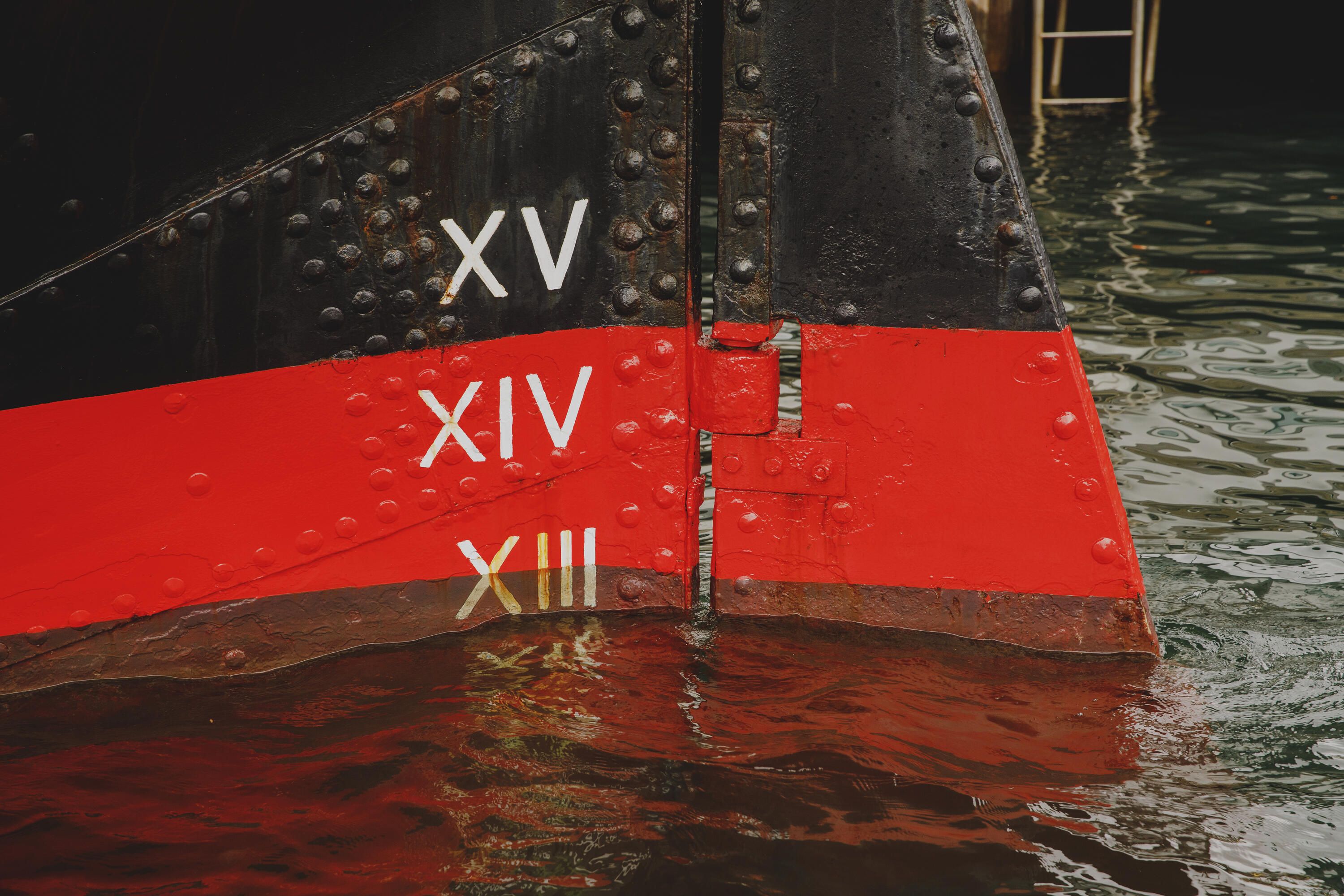 Close up photo of a black and red ship's hull in the water with roman numerals painted in white.