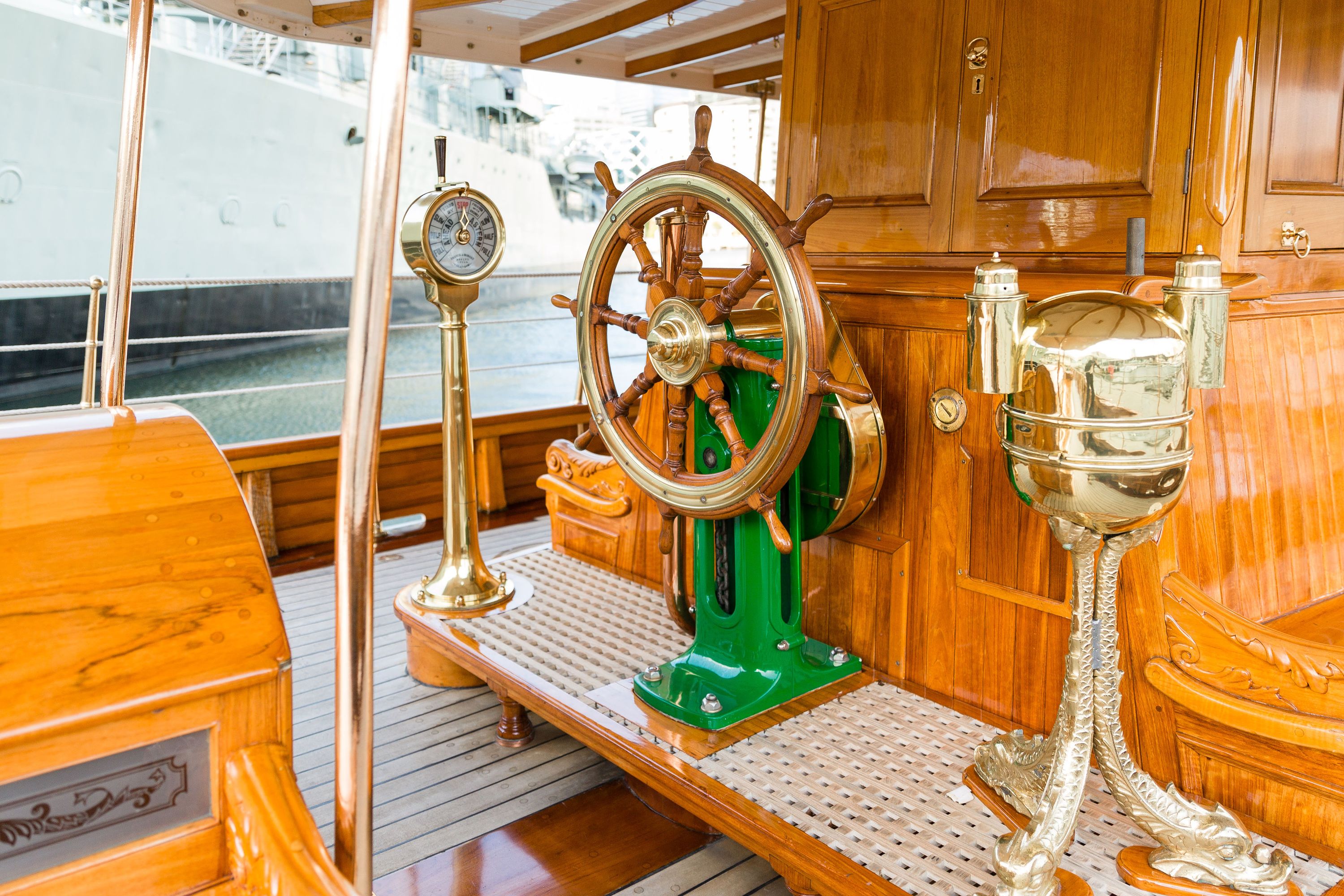 Photo showing detail of a wooden steam yacht, showing the wheel with brass telegraph on the left and brass binnacle on the right.