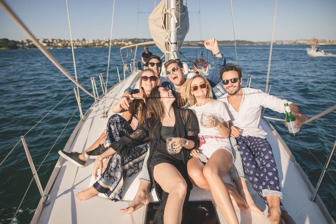 A group of people smiling and posing for a group photo in the sun, on the top deck of a yacht. Sydney harbour is behind them.