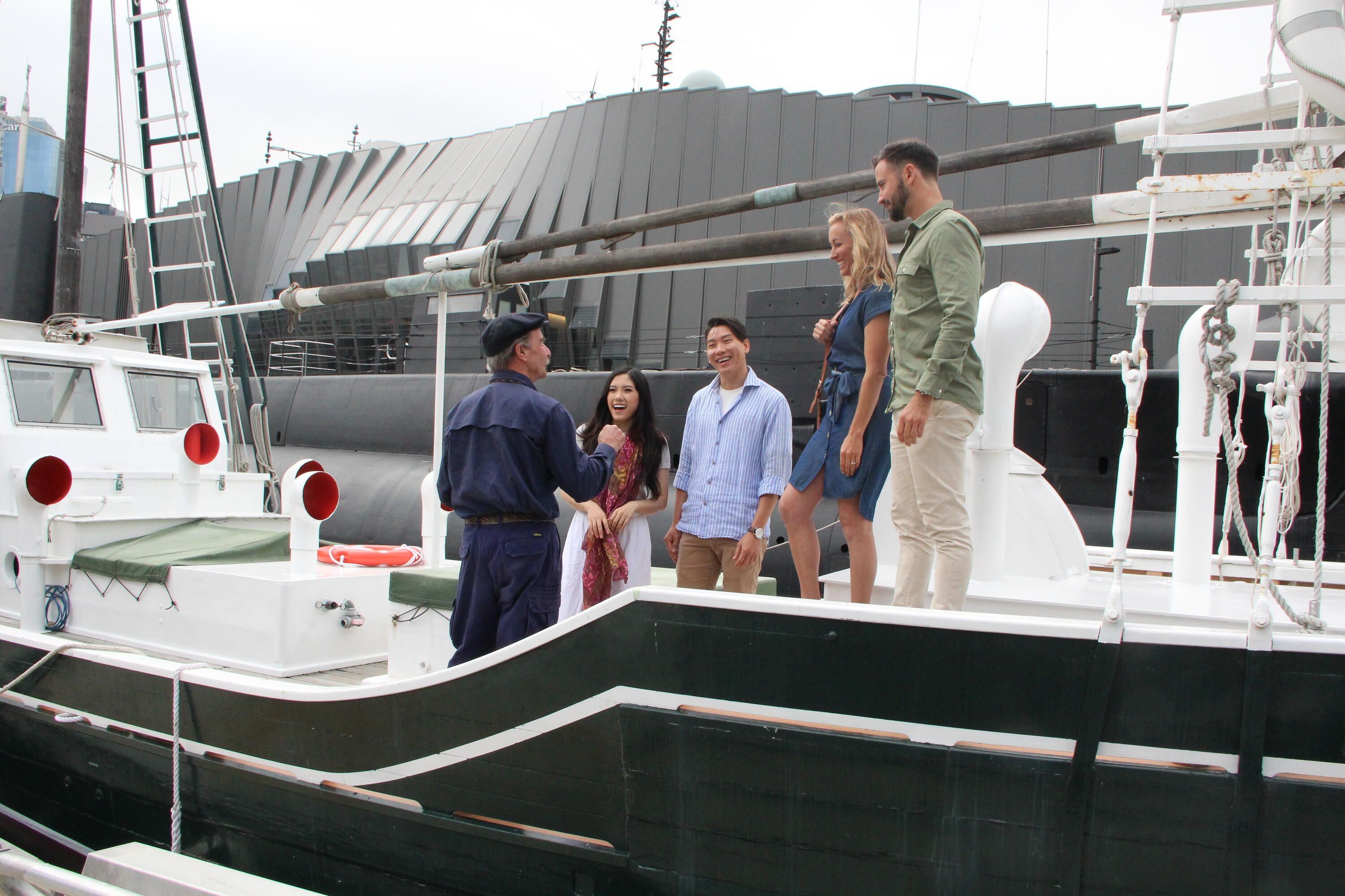 Photo of a volunteer in blue uniform talks to a small group of adult visitors onboard a white and green boat.