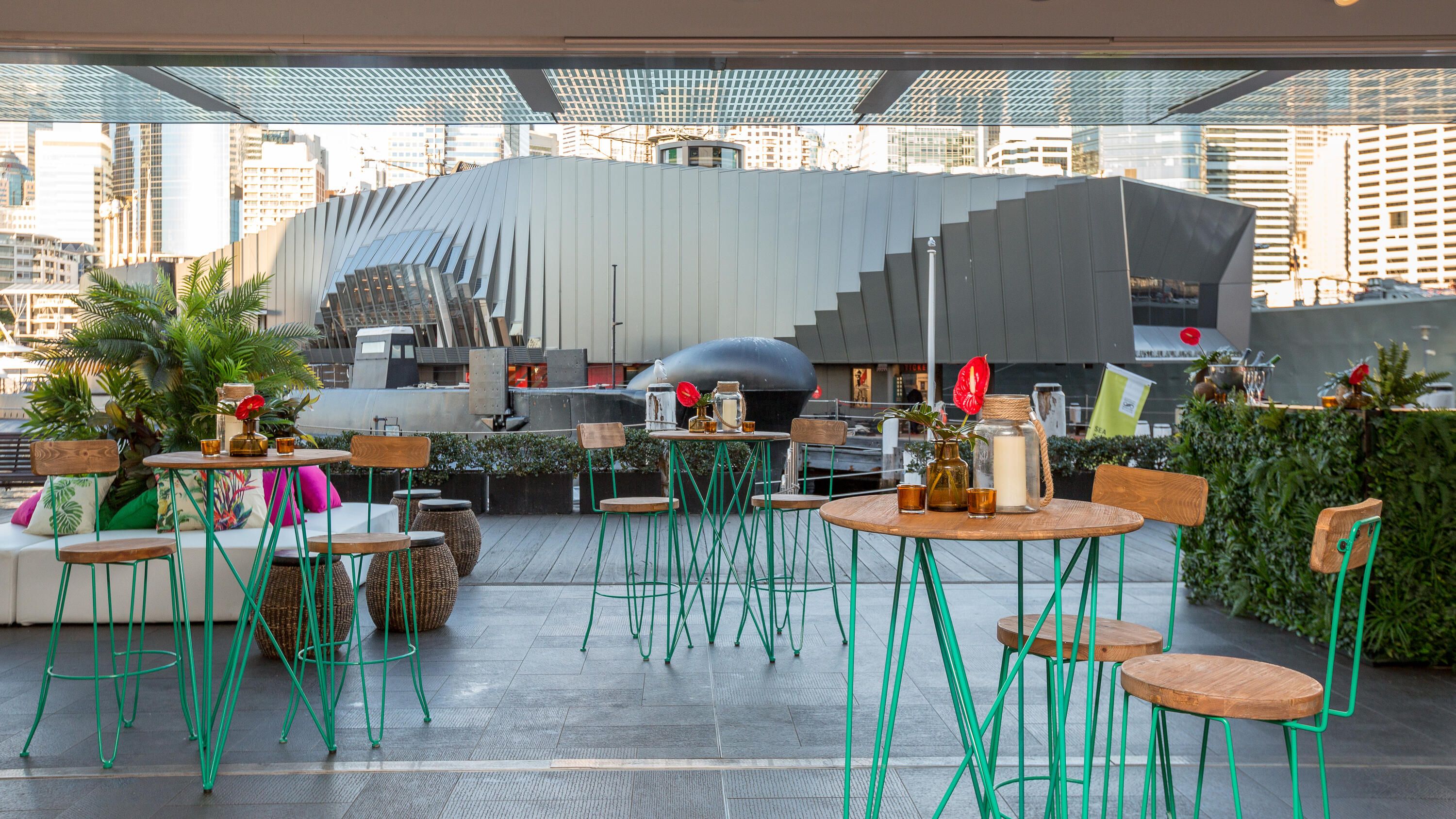 Photo from an interior opening into an exterior space, with natural and teal coloured event furniture and a submarine in the background.