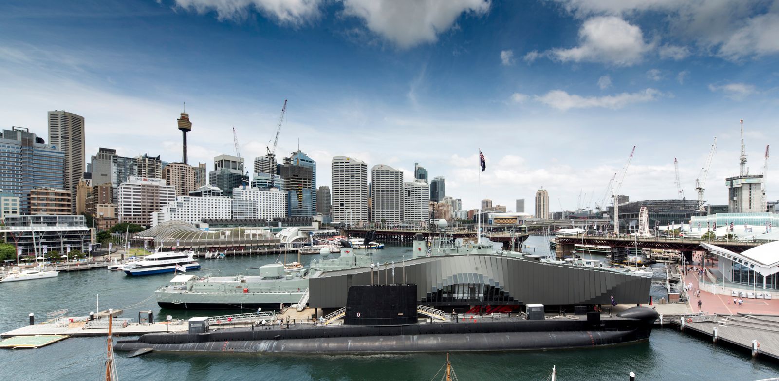 This wide elevated exterior view showcases both the building's unique architecture as well as its location in relation to the city and Darling Harbour precinct. The ONSLOW submarine, shown in profile, features prominently in front of the building. The Pyrmont Bridge and city skyline appear in the background. 