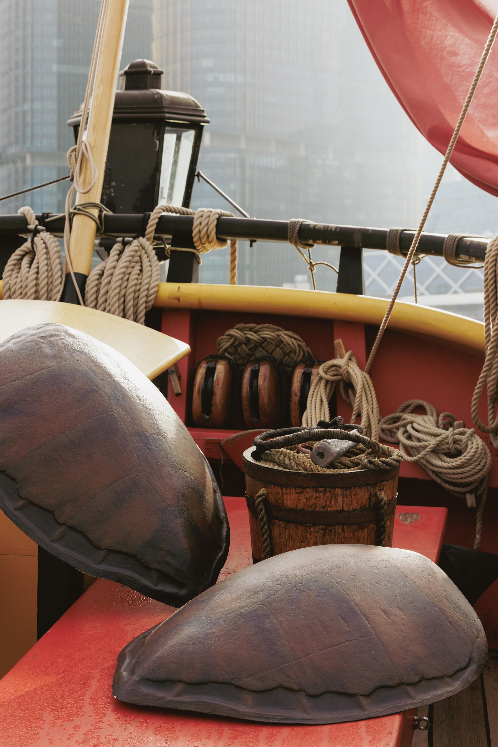 A close up photo showing 2 large turtle shells and a wooden bucket. Behind them can be seen the back rail of a tall ship, a lantern and the edge of a red flag. 