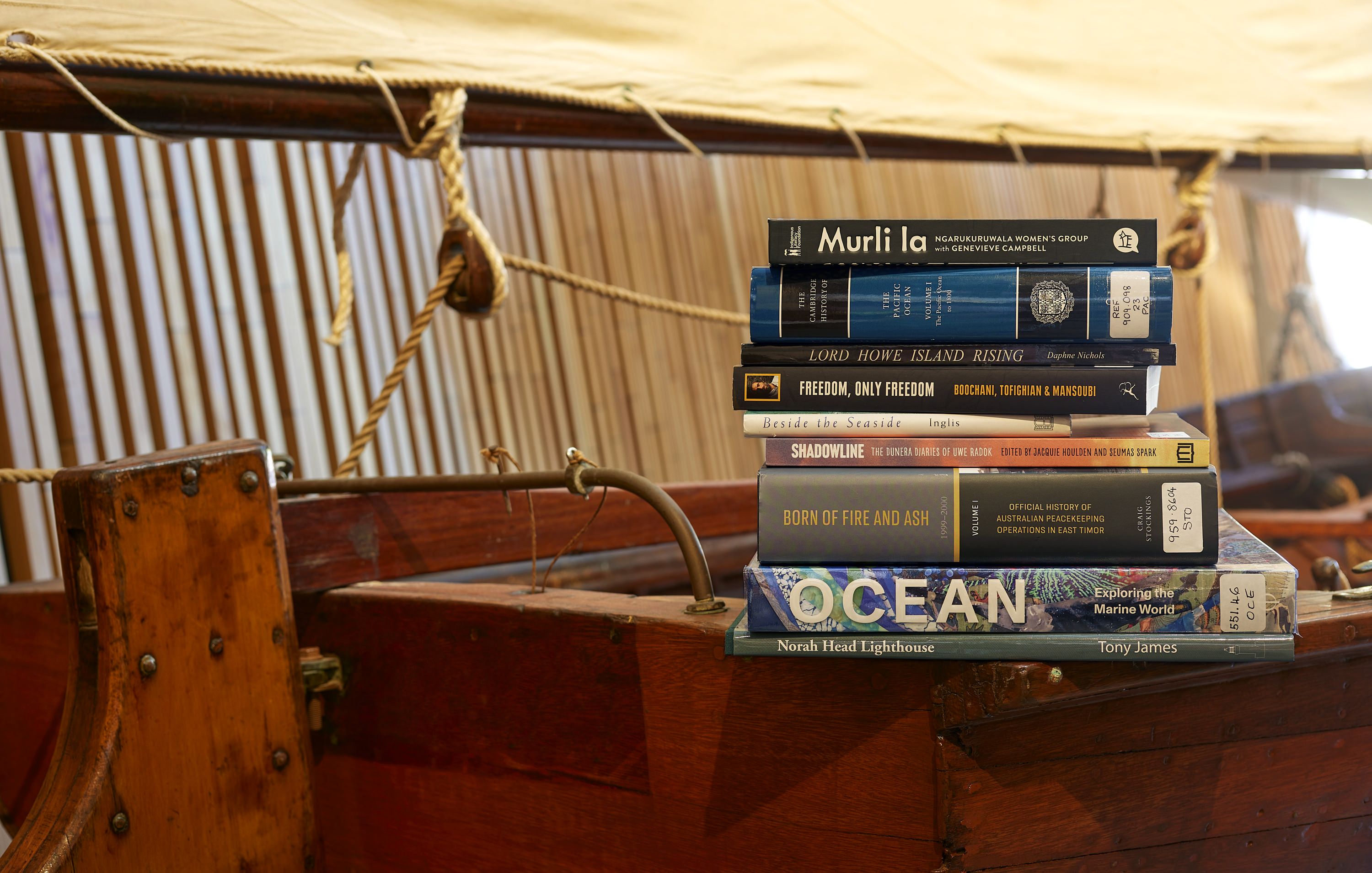Pile of library books with different coloured covers sitting on the side of a wooden boat. 