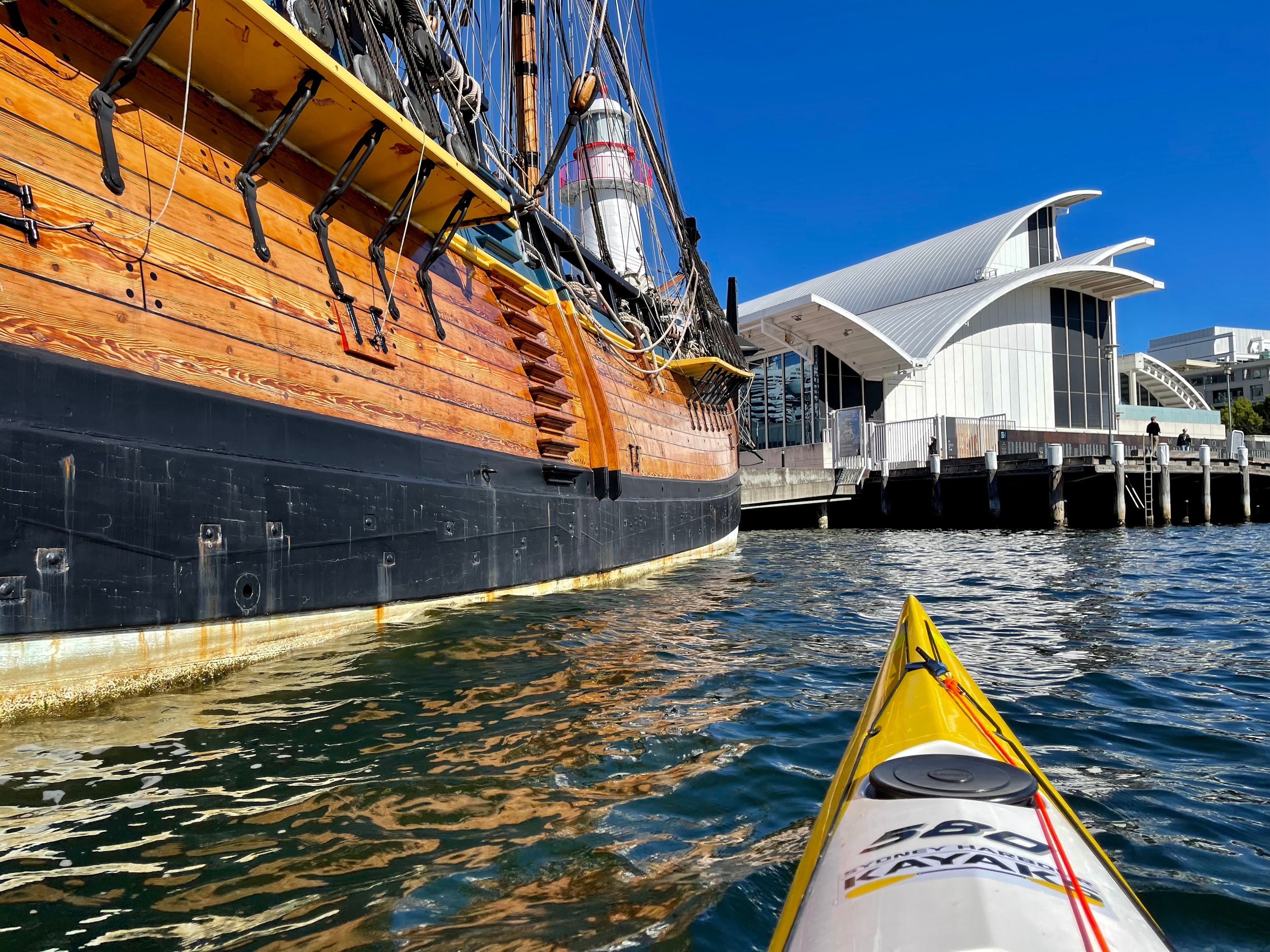 Photo taken from a kayak on the water, looking up at the museum building and the side of a wooden tall ship.