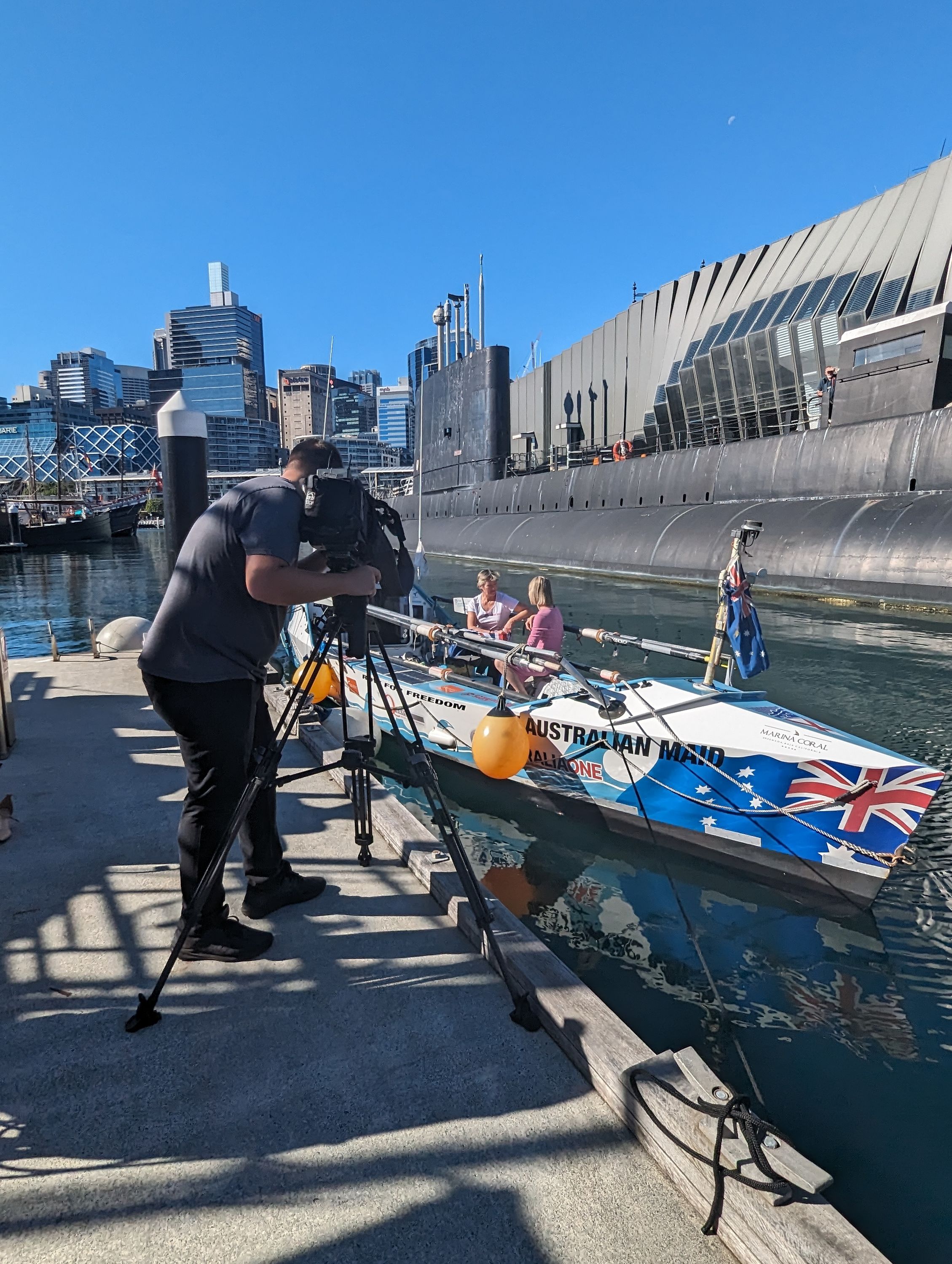 photo showing behind the scenes, with a camera man standing on the wharf, taking video of two ladies in a boat.