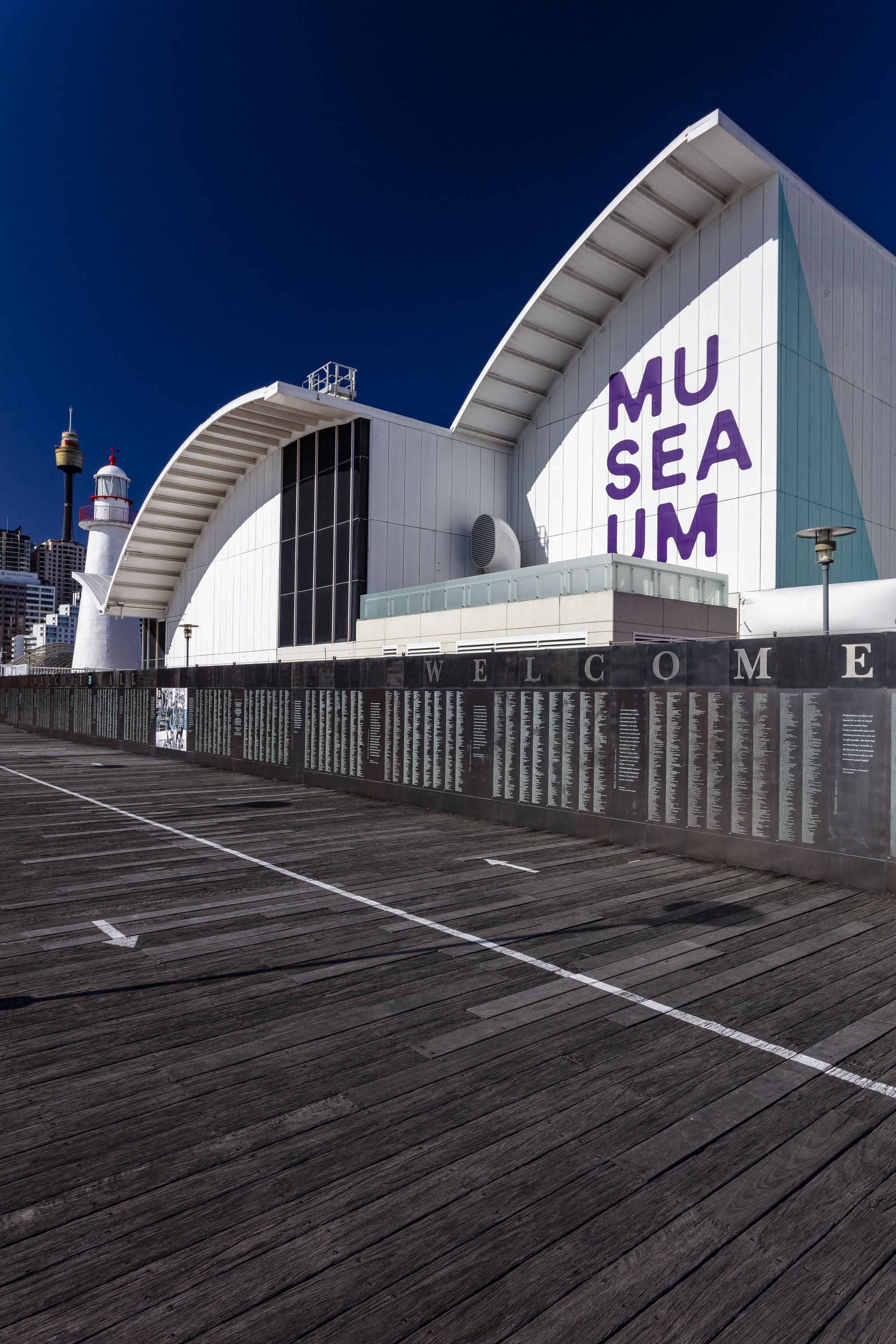 Photo showing a dark, metal wall covered with small, green engraved names in front of a large white building featuring the museum logo. 