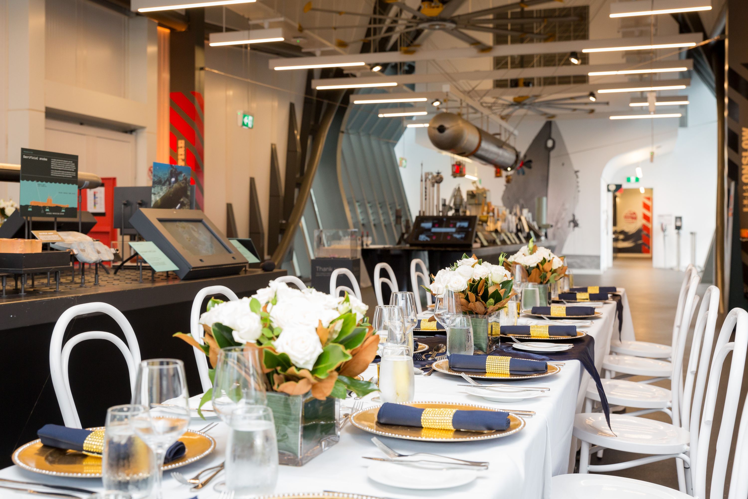 photo of a long table with white tablecloth set for a formal event and decorated with flowers, in a museum space.