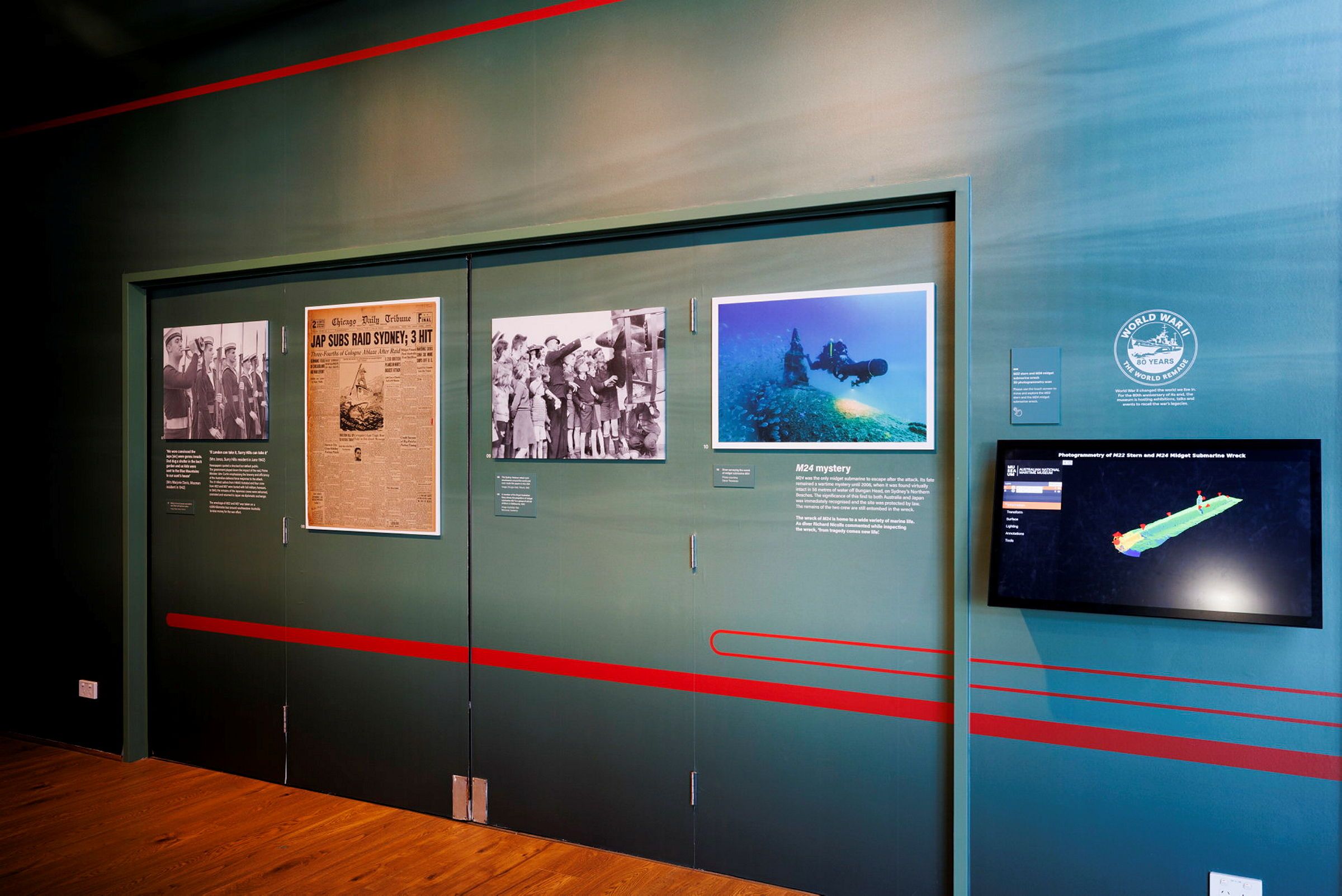Photo showing the wall of an exhibition space, with large images from newspapers and of underwater divers. There is also a screen with a 3D model of a sunken submarine. 