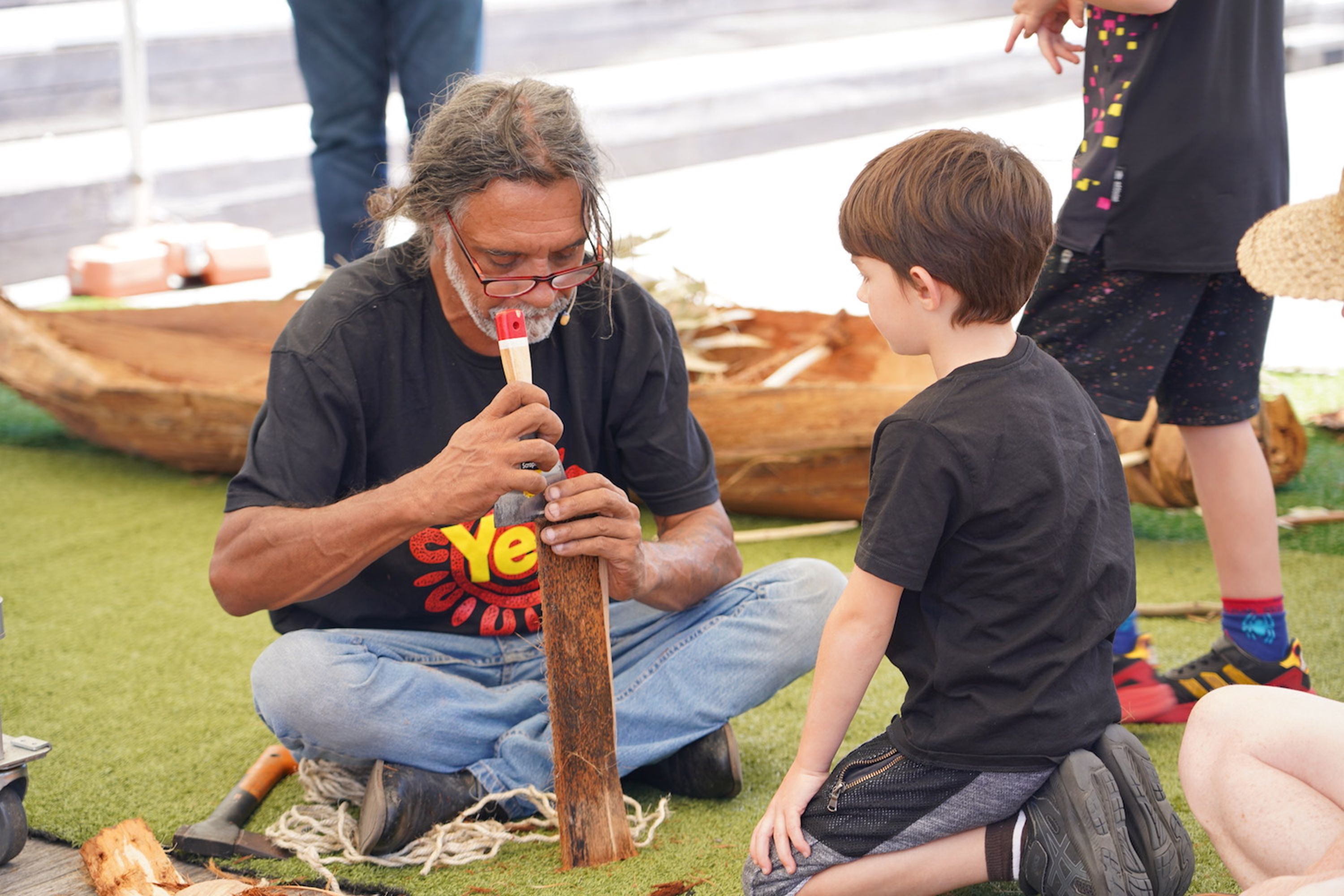 Man and a boy kneeling on the ground. The man us using a tool to work on a piece of bark.