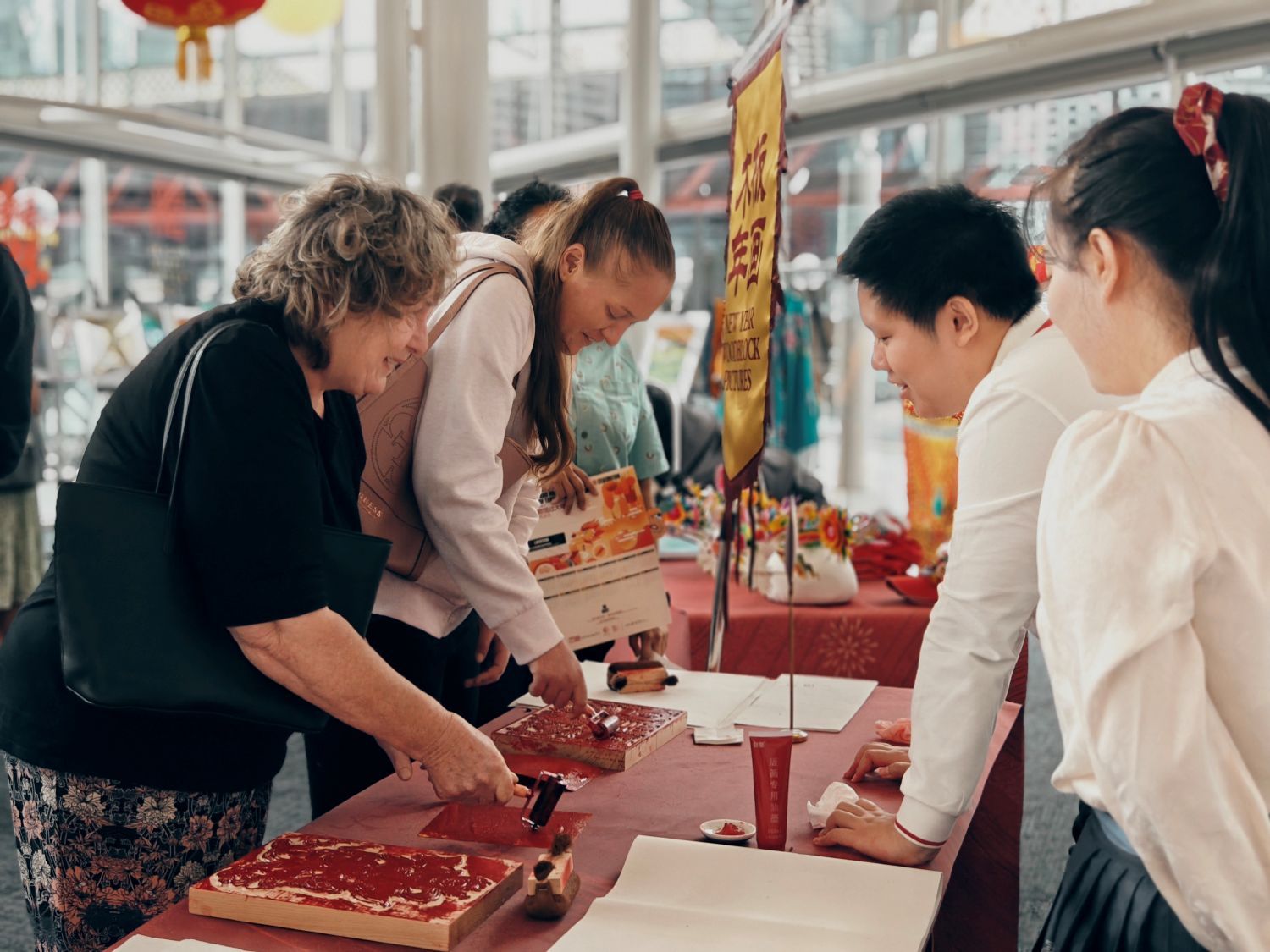 Photo with 2 instructors wearing white shirts on the right, instructing two women on the left how to make a roll ink onto a wood block to make a print.