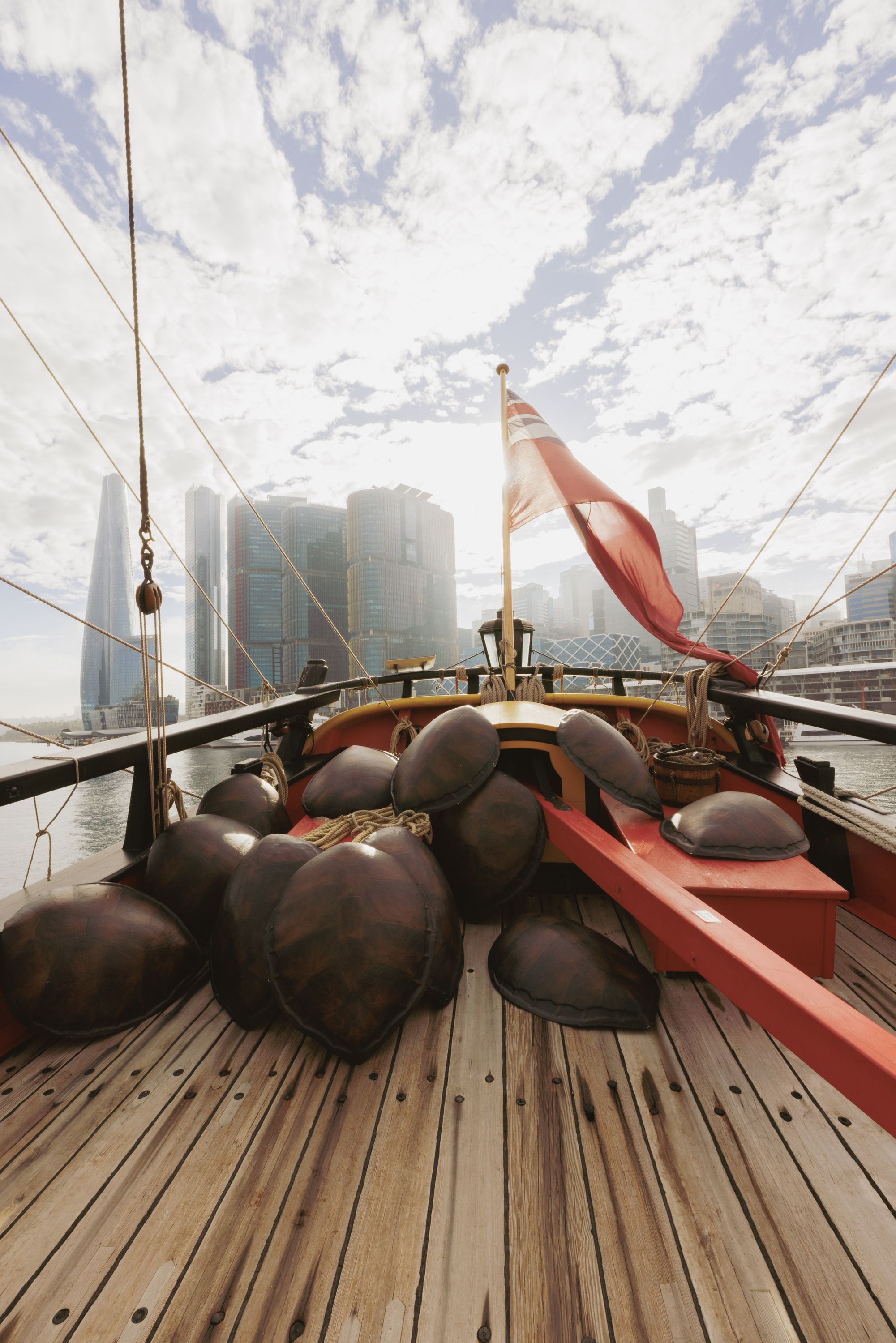 Photo showing the back of a wooden tall ship with a collection of turtle shells on the deck. There is a red flag flying on the ship, and city buildings in the background. 