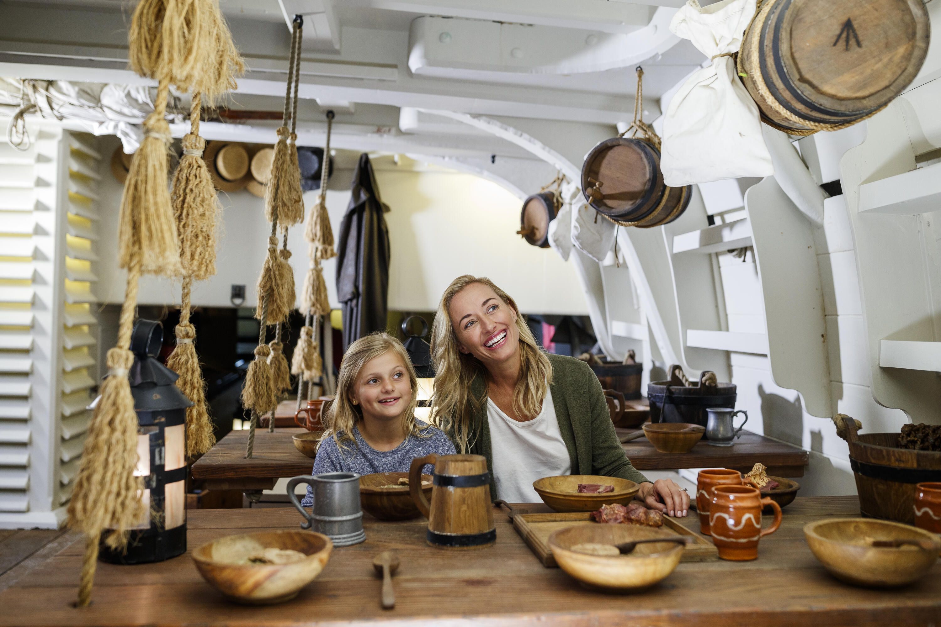 Photo taken inside a wooden tall ship showing a mother and daughter sit at a table with an assortment of tankards and bowls.
